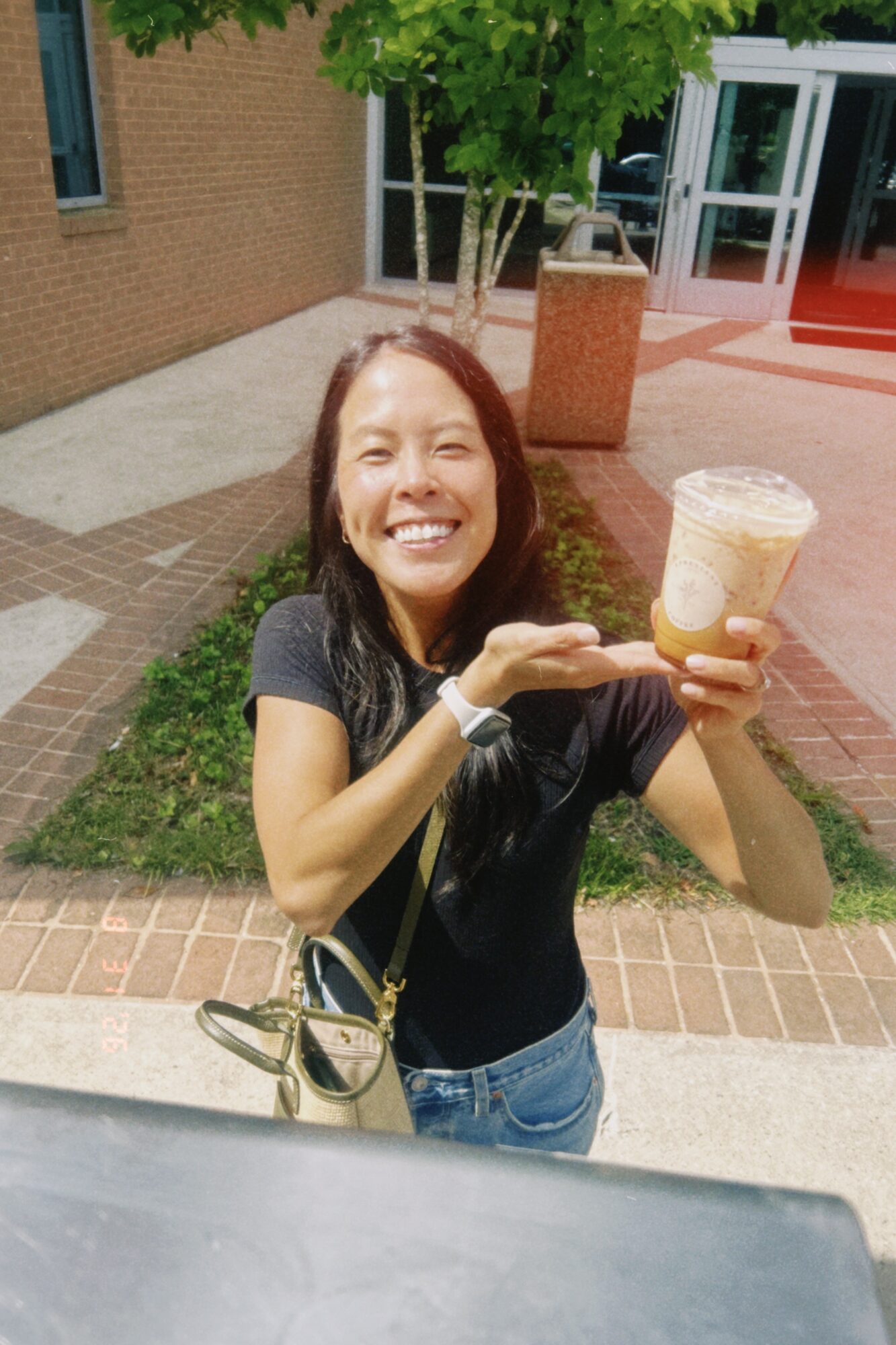 Woman smiling outdoors holding a large iced beverage, standing on brick sidewalk near a tree and building entrance.