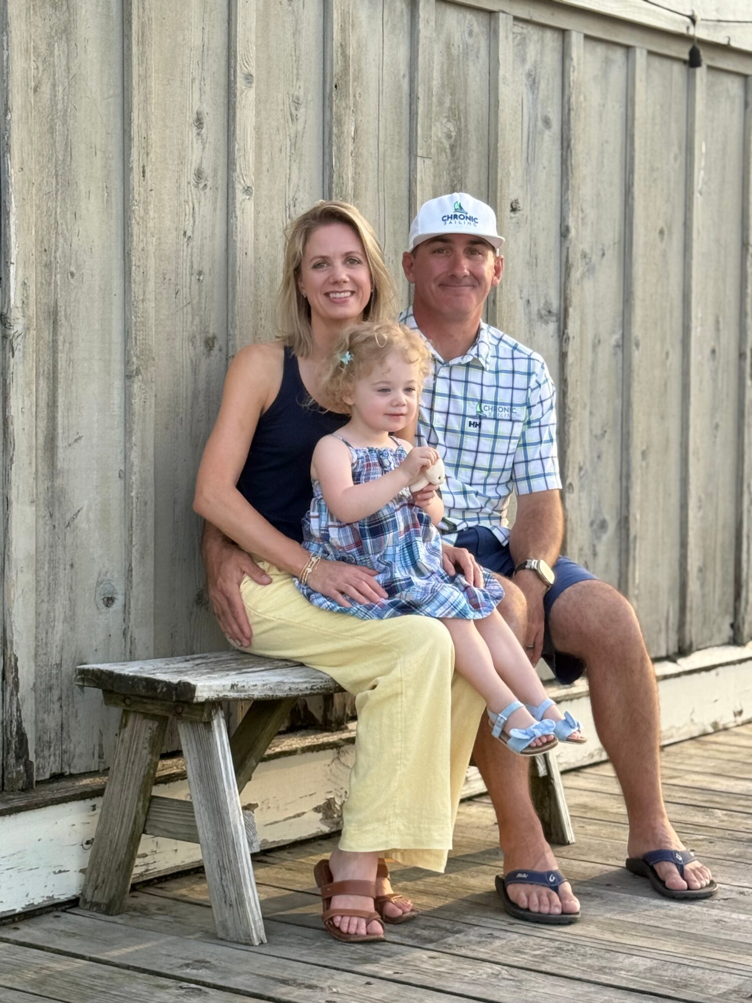 Family of three sitting on a wooden bench outdoors, smiling, with a wooden fence in the background.
