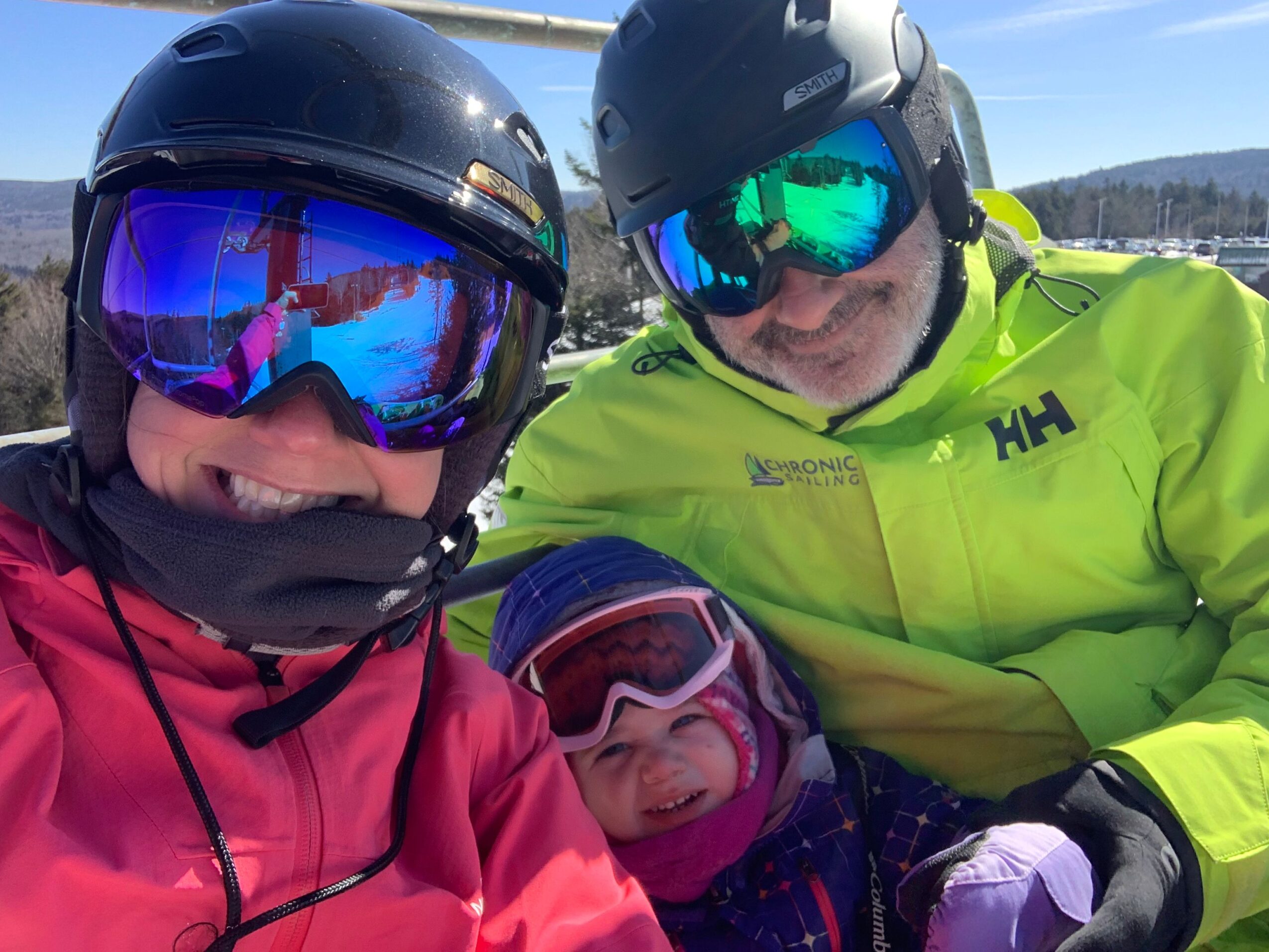 Three people in winter gear, wearing helmets and goggles, lying on snow with a blue sky background.