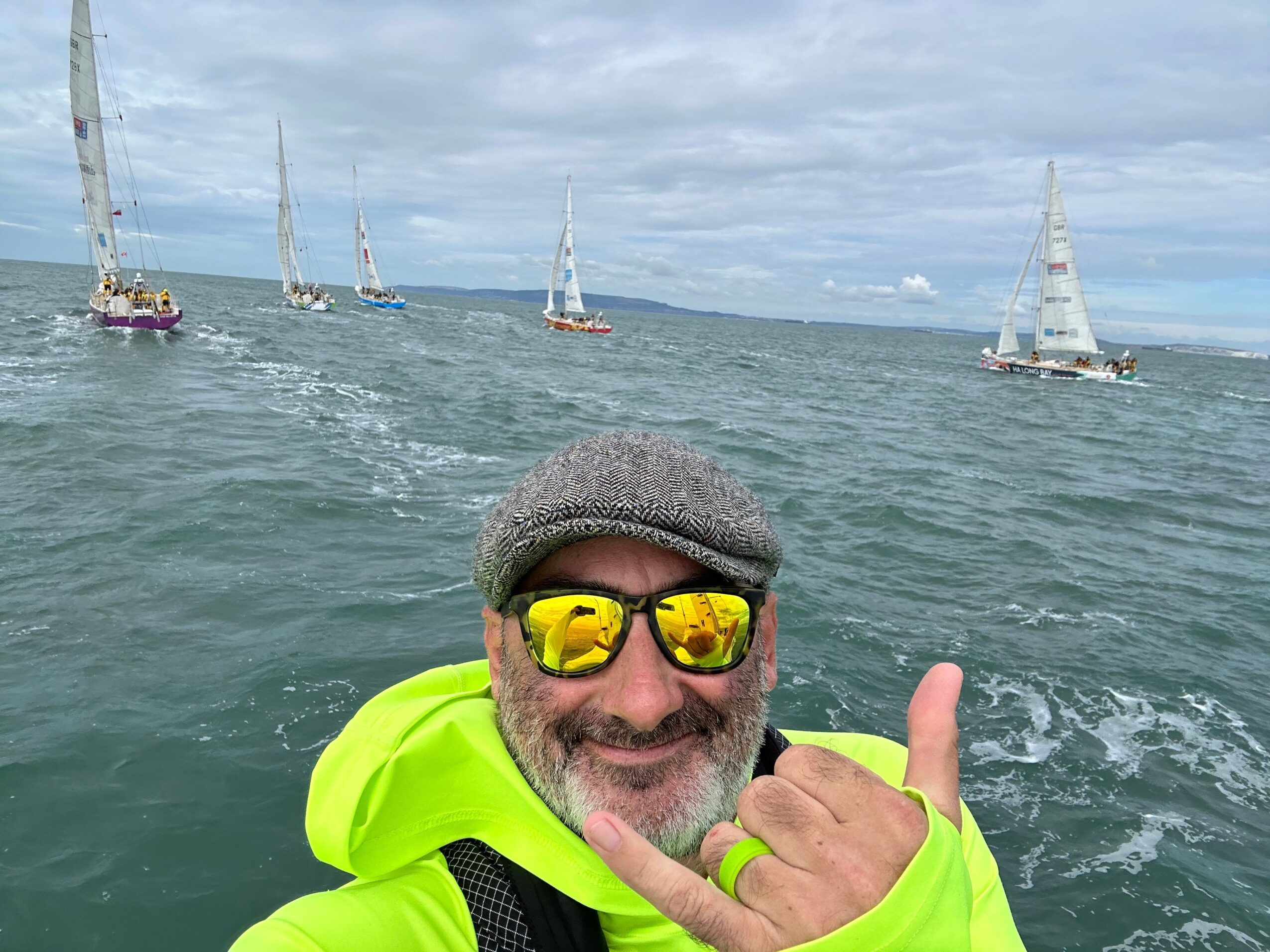 Man wearing sunglasses and a bright yellow jacket, upside down in water, with a cloudy sky and wind turbines in background.
