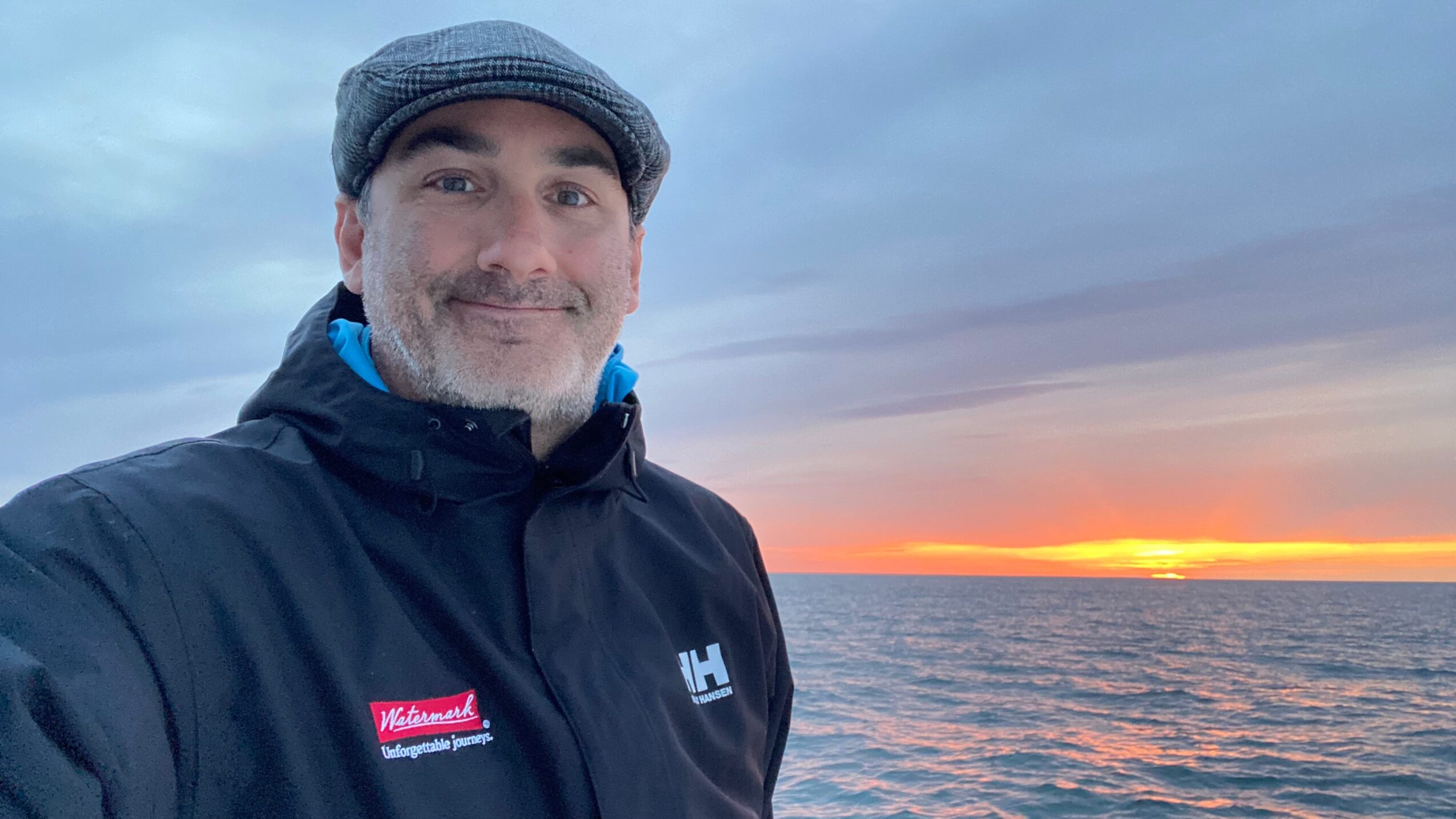 Man wearing a jacket and cap standing near water during sunset, smiling at the camera.