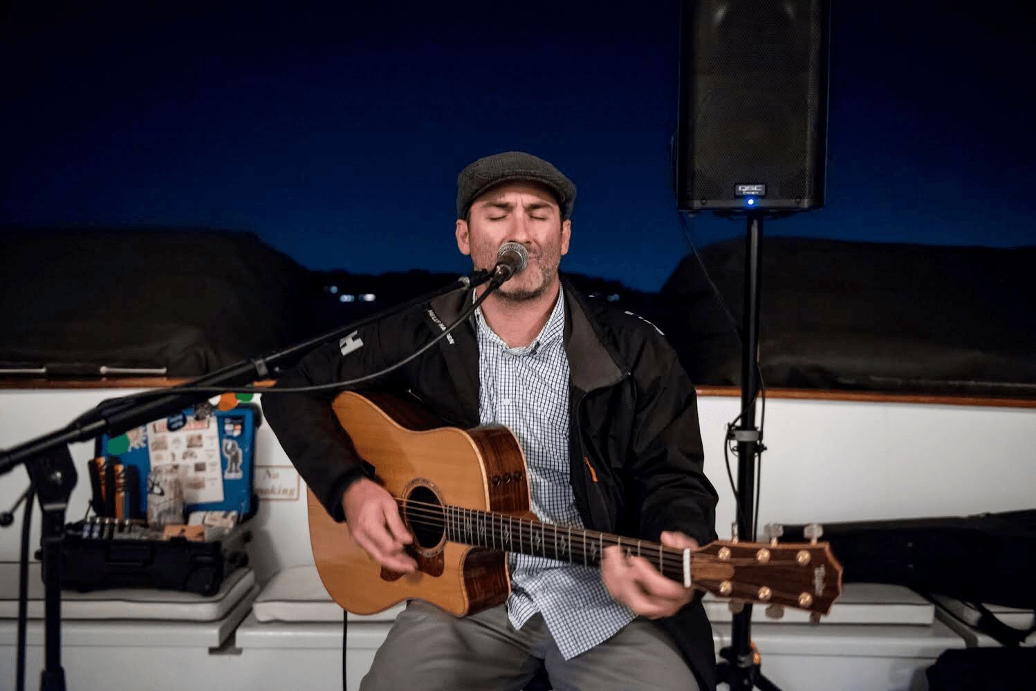 Man playing acoustic guitar and singing into microphone outdoors at night, with a speaker and equipment nearby.