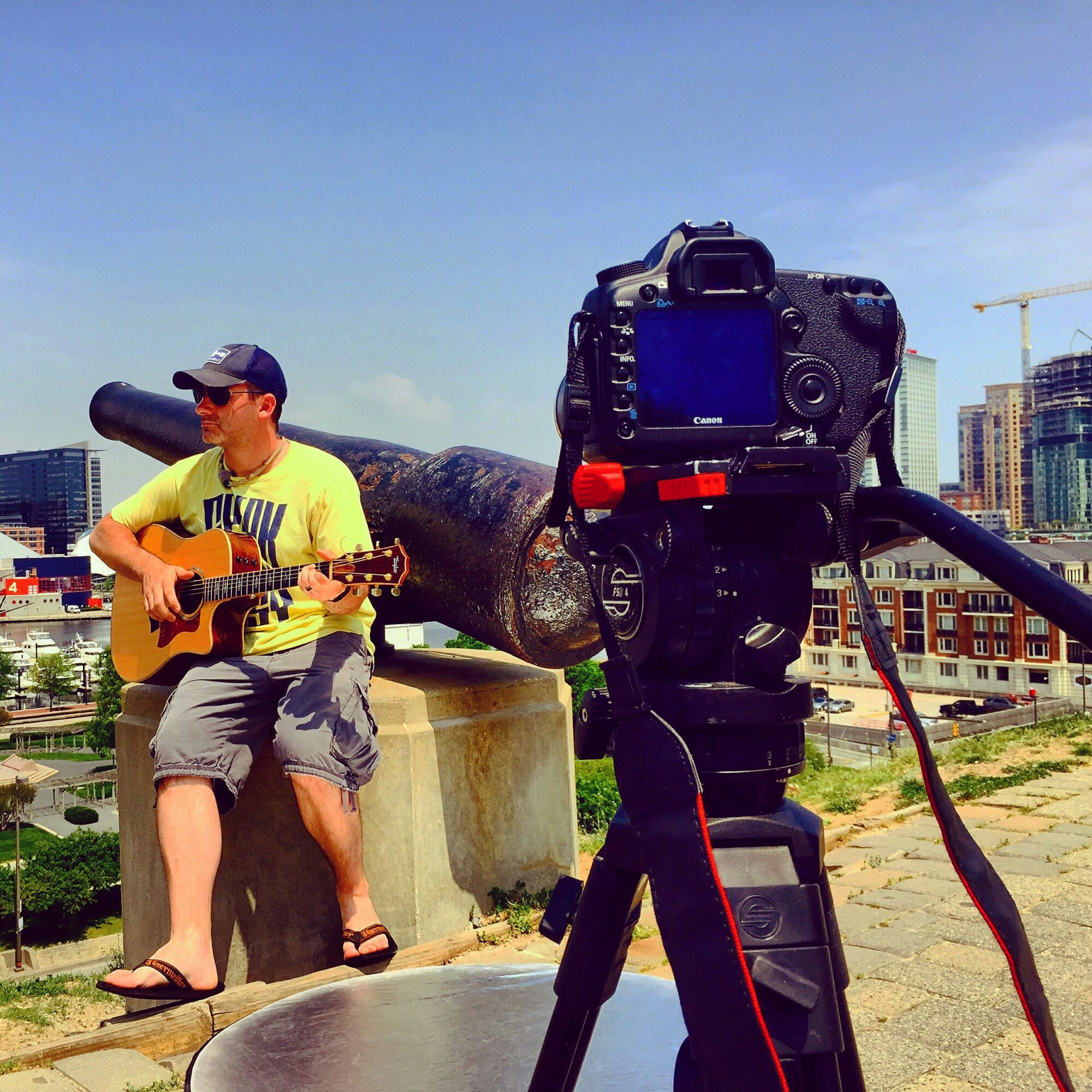Person playing guitar outdoors with camera on tripod capturing the scene, city buildings in background, sunny day.