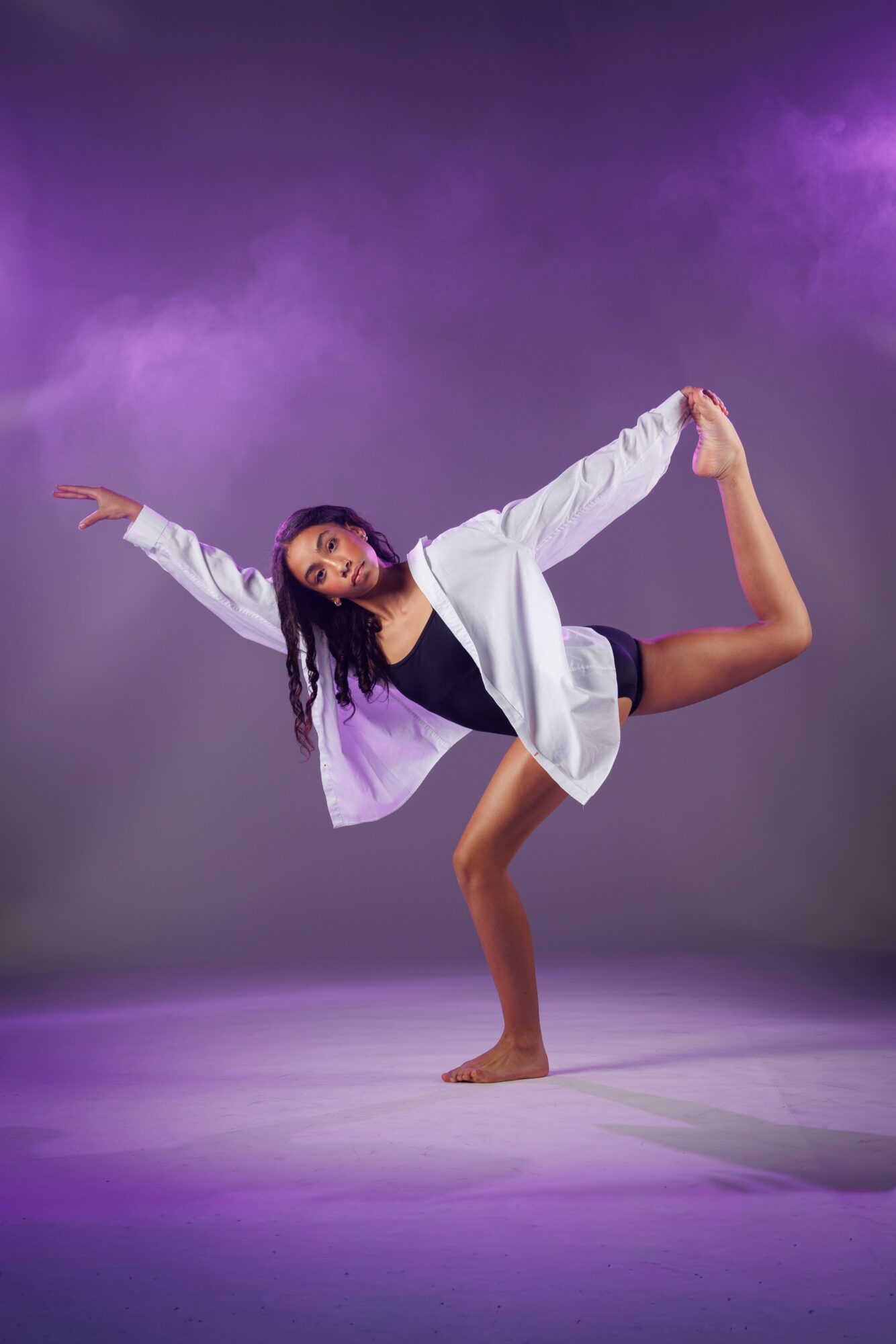 A dancer balances on one leg with arms extended, wearing a white jacket and black shorts, against a purple background.