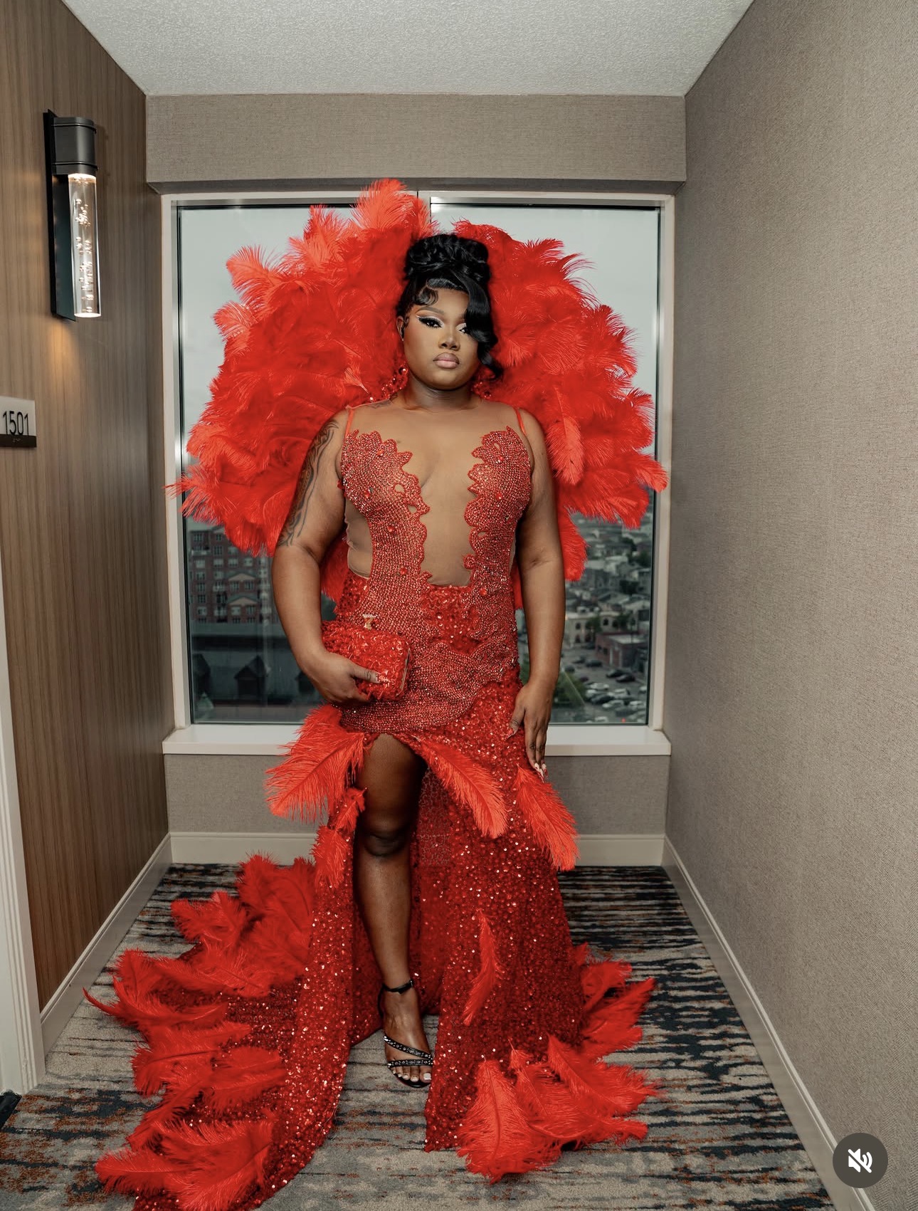 Woman in red dress with large feathered backpiece, standing in room with window, patterned carpet, and neutral walls.