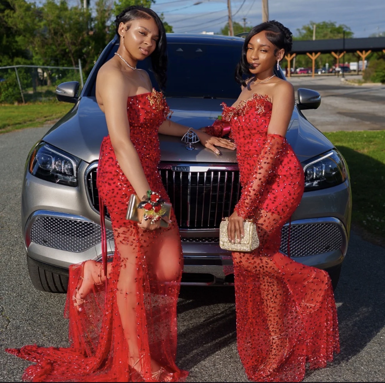 Two women in red dresses stand in front of a silver car outdoors, holding handbags and posing.