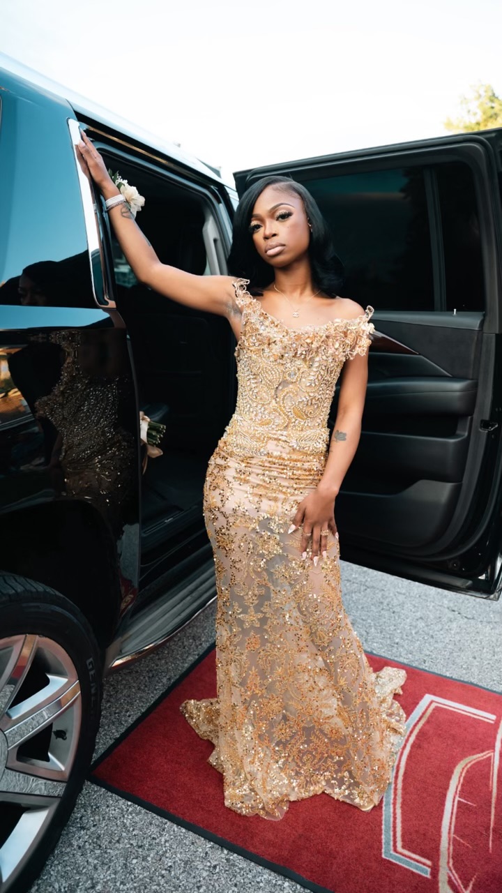 Young woman in a gold, sequined gown standing outside a black vehicle, holding the door open with one hand, looking at the camera.