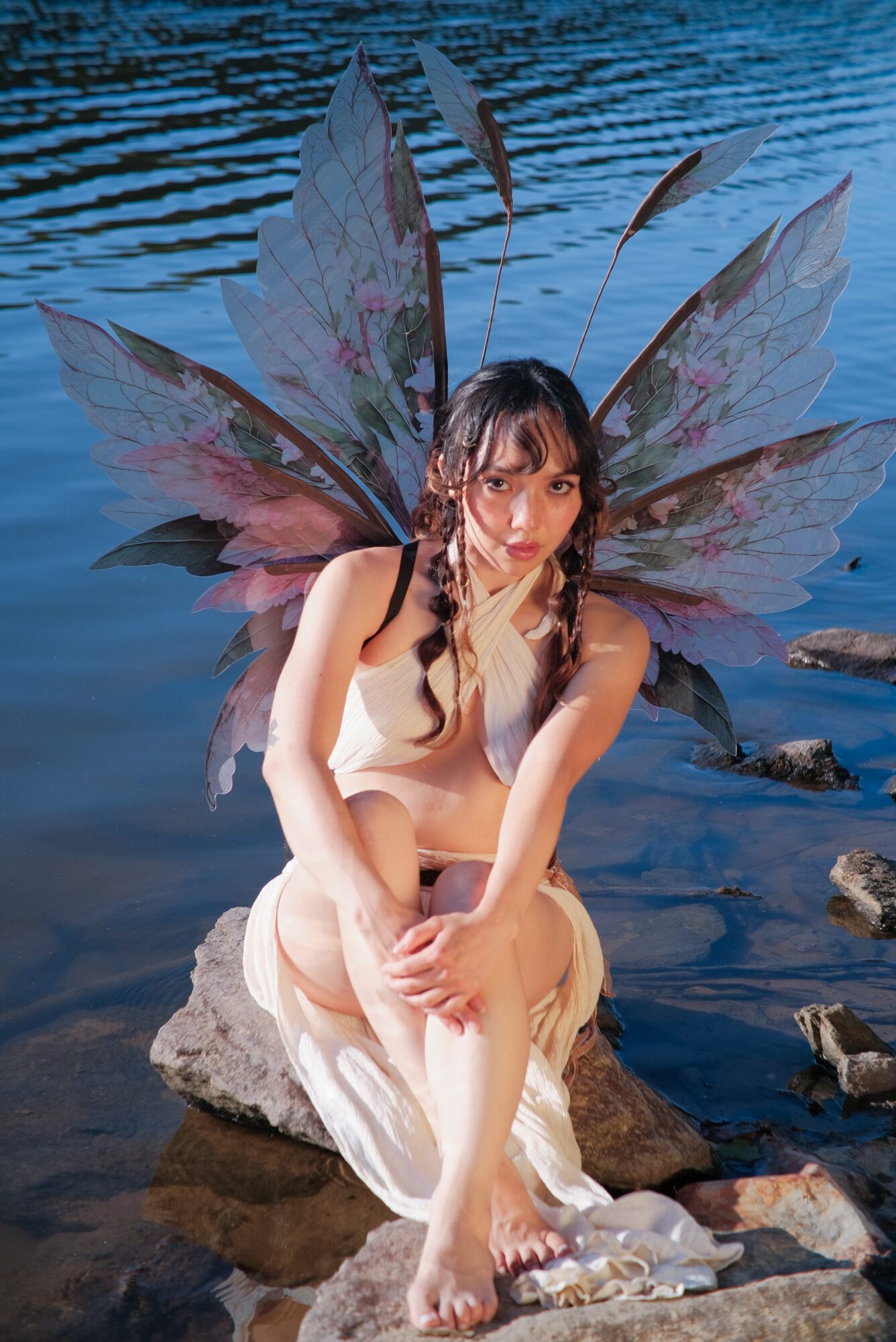 Young woman with fairy wings sitting on rocks by water, looking thoughtful, wearing a light dress.