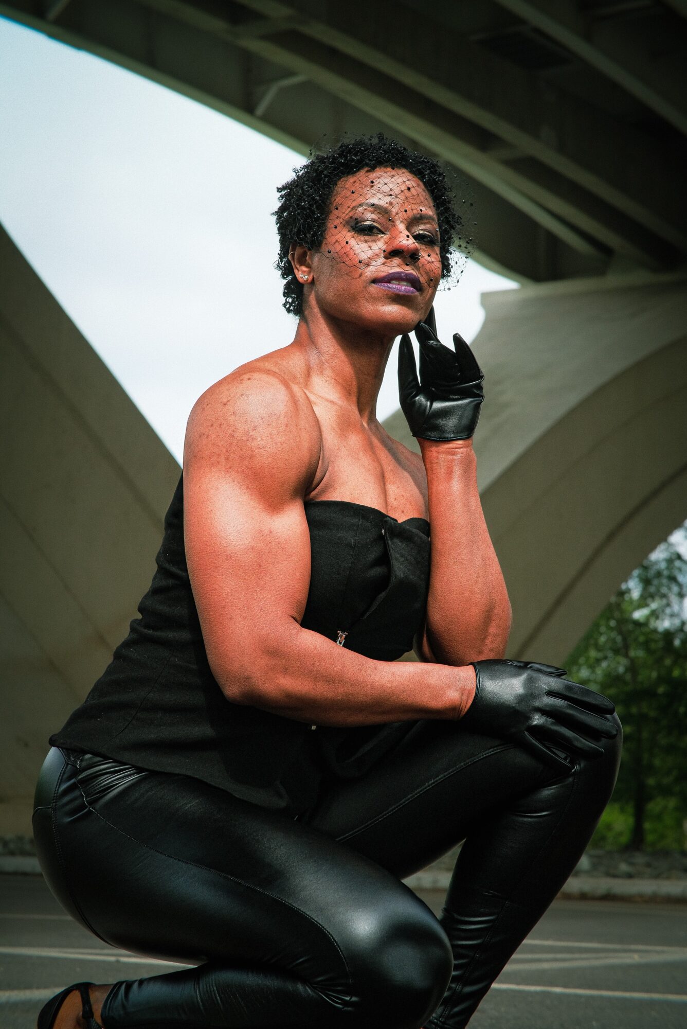 Woman with short curly hair and glitter makeup, wearing black strapless top and leather pants, squatting outdoors under a bridge, touching her face with gloved hand.