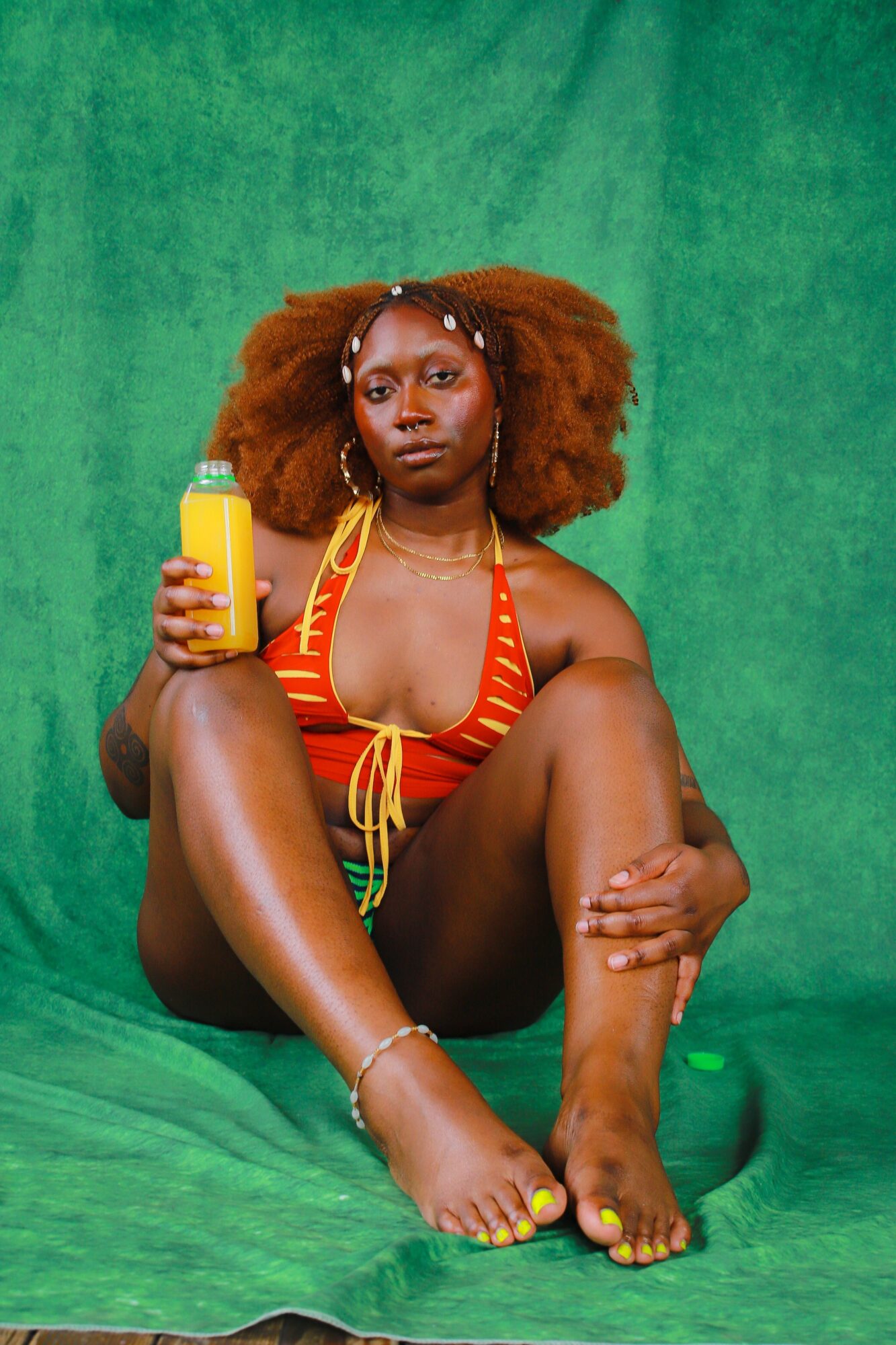 Woman with curly hair in a red and yellow bikini sitting on a green background, holding a yellow bottle.