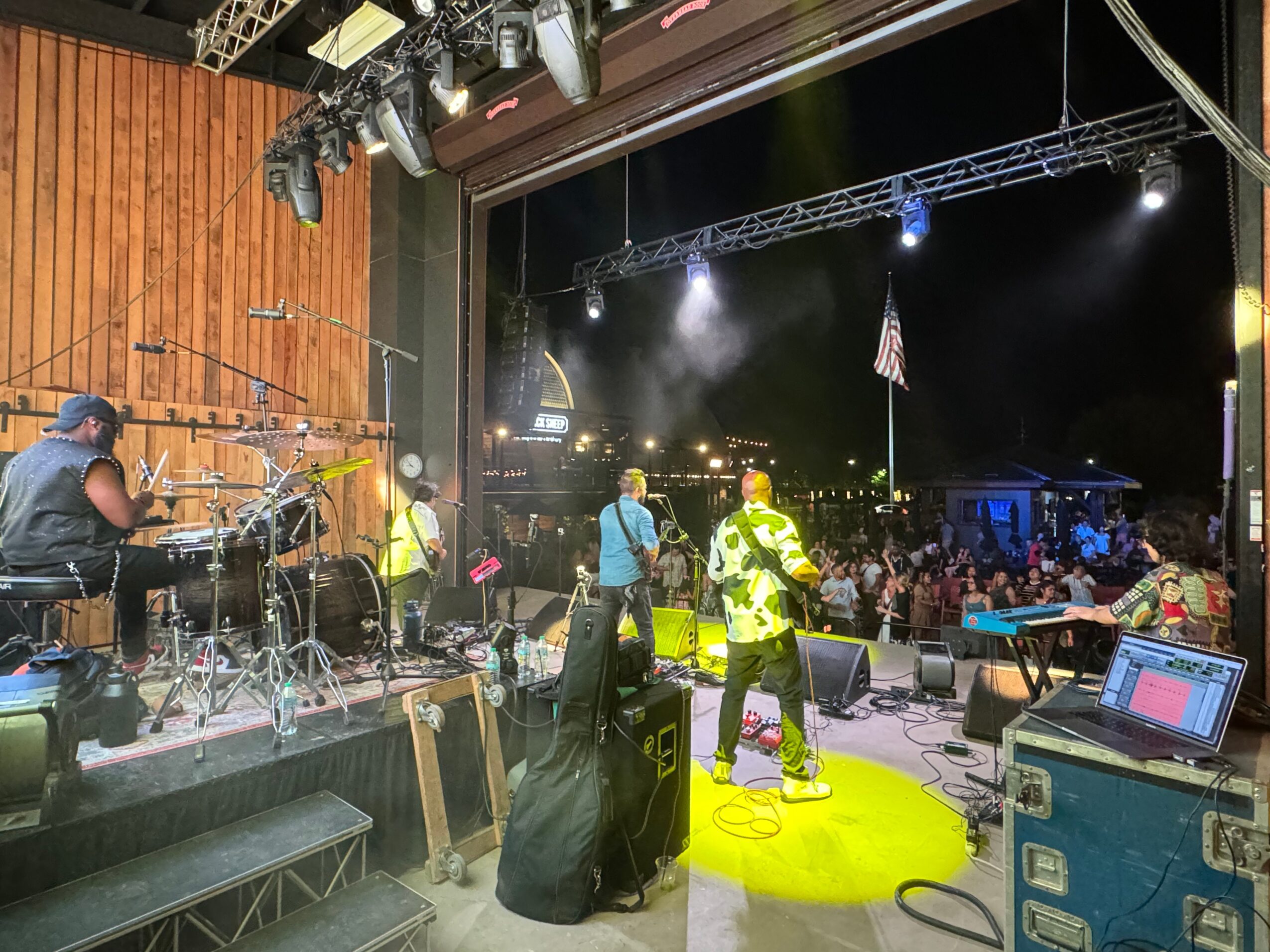 Musicians perform on stage with an audience in front, stage lights, and an American flag in the background.