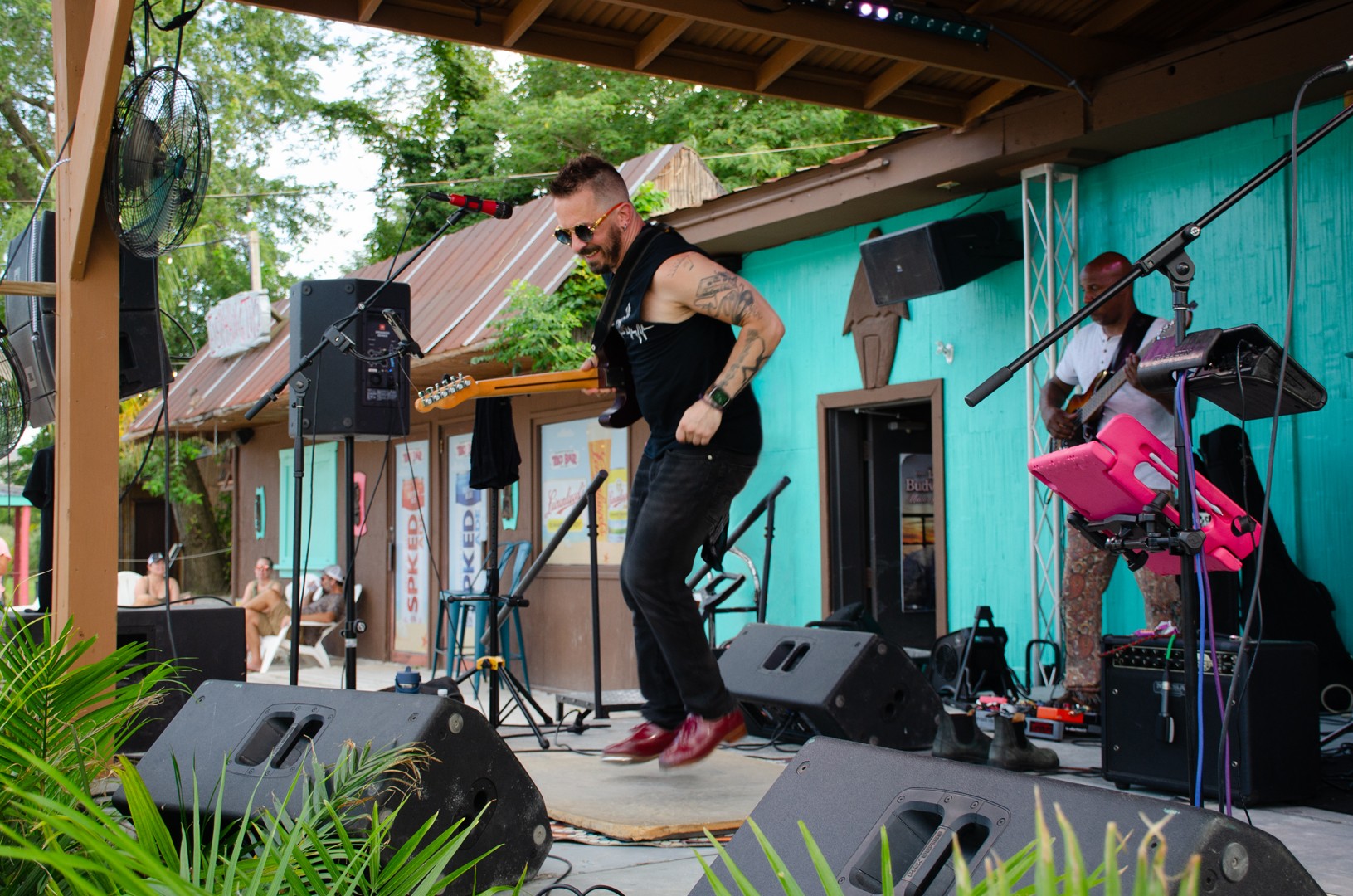 Man dancing on stage with musical equipment, outdoors, green trees in background, colorful building behind, smiling, wearing black vest and red shoes.