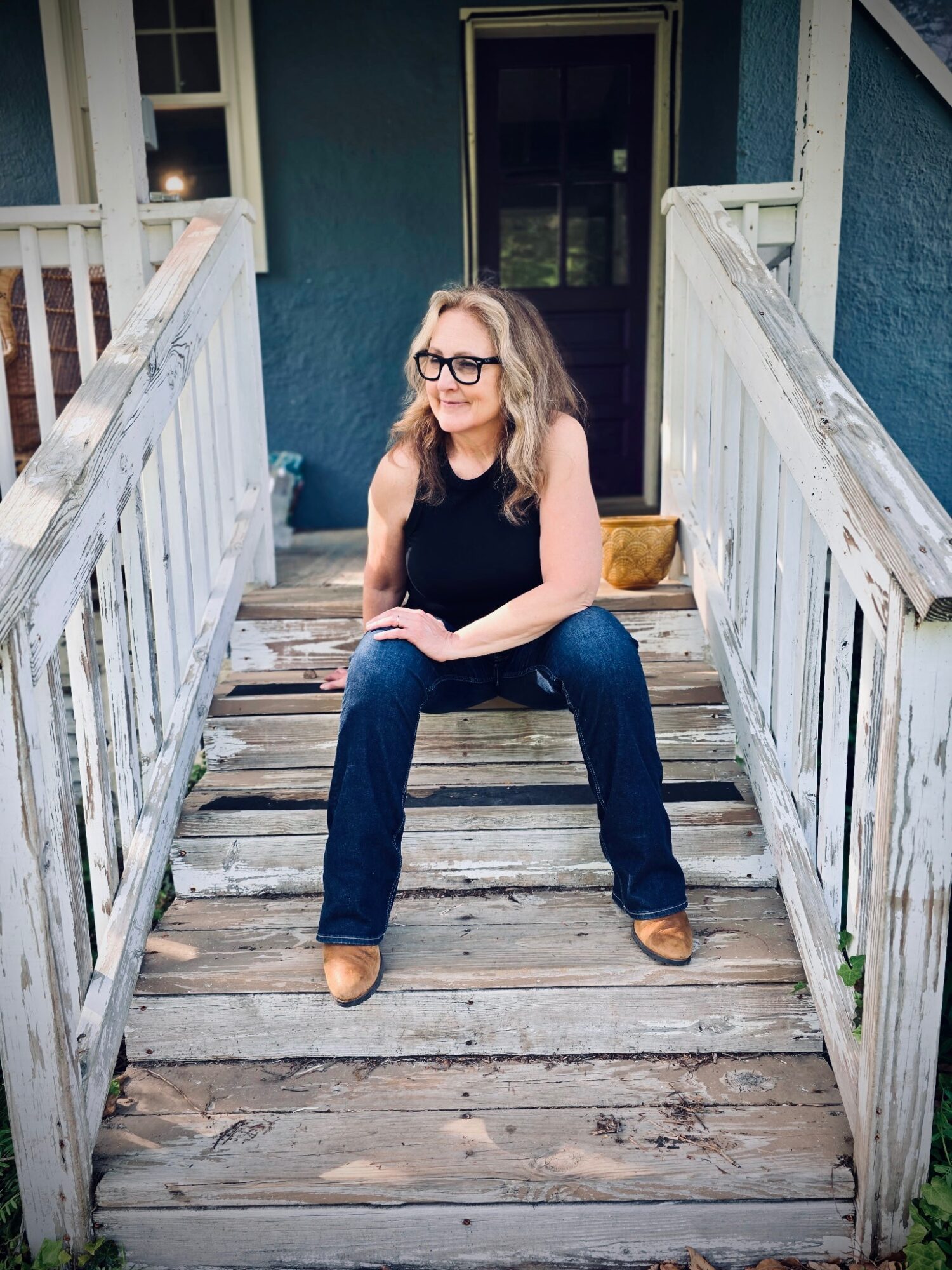 Woman with glasses sitting on wooden stairs outside a house, smiling, wearing a black top and jeans.