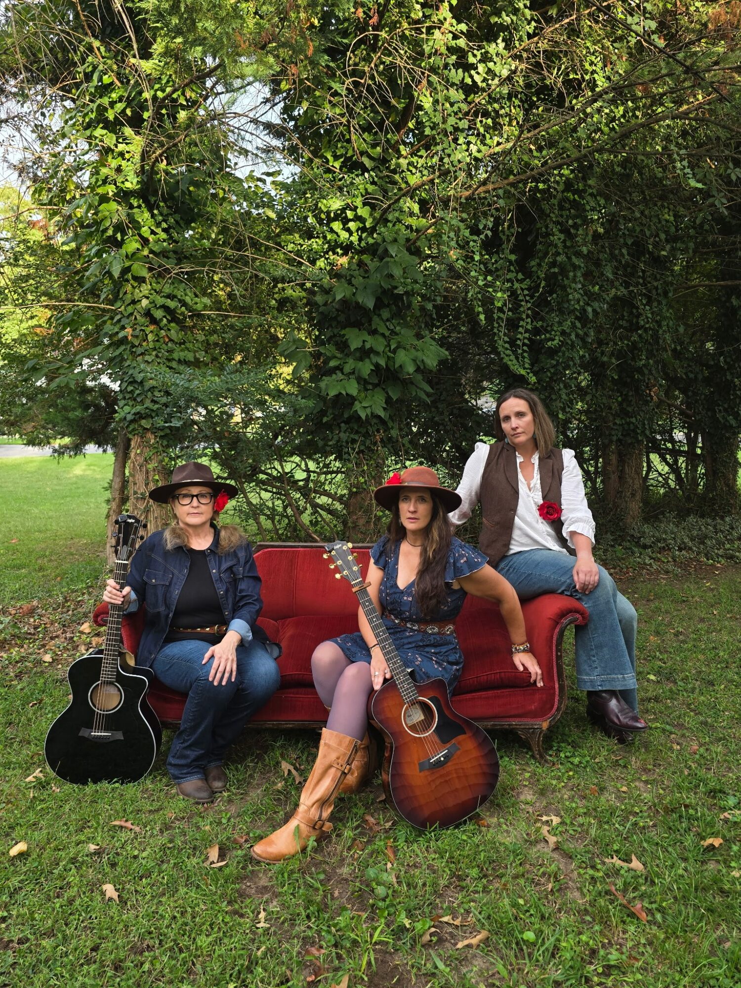 Three women outdoors with trees, two sitting on a red sofa, one standing behind, two holding guitars, all wearing hats.