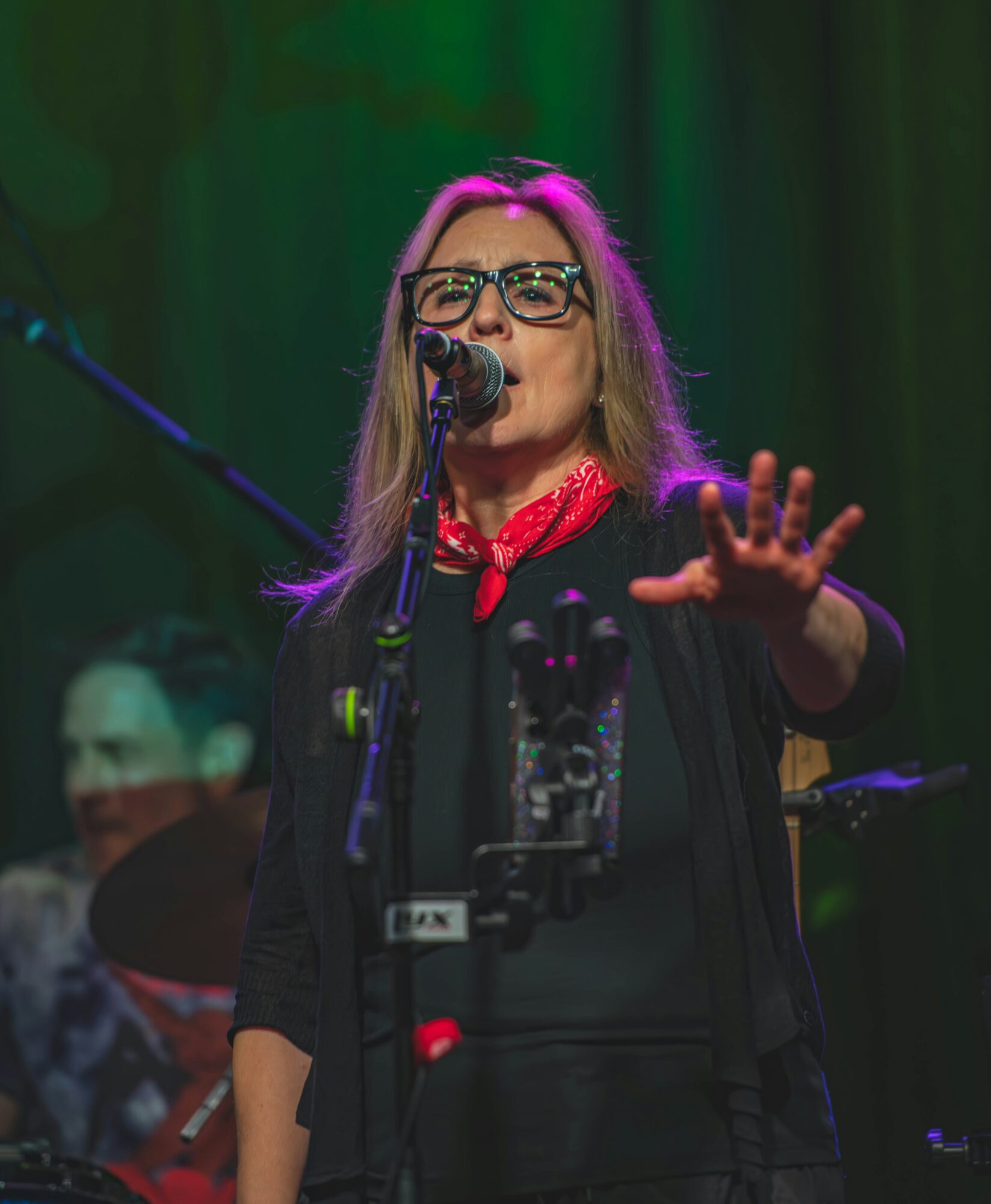 Woman with glasses singing into microphone on stage, hand raised, green curtain background, colorful lighting