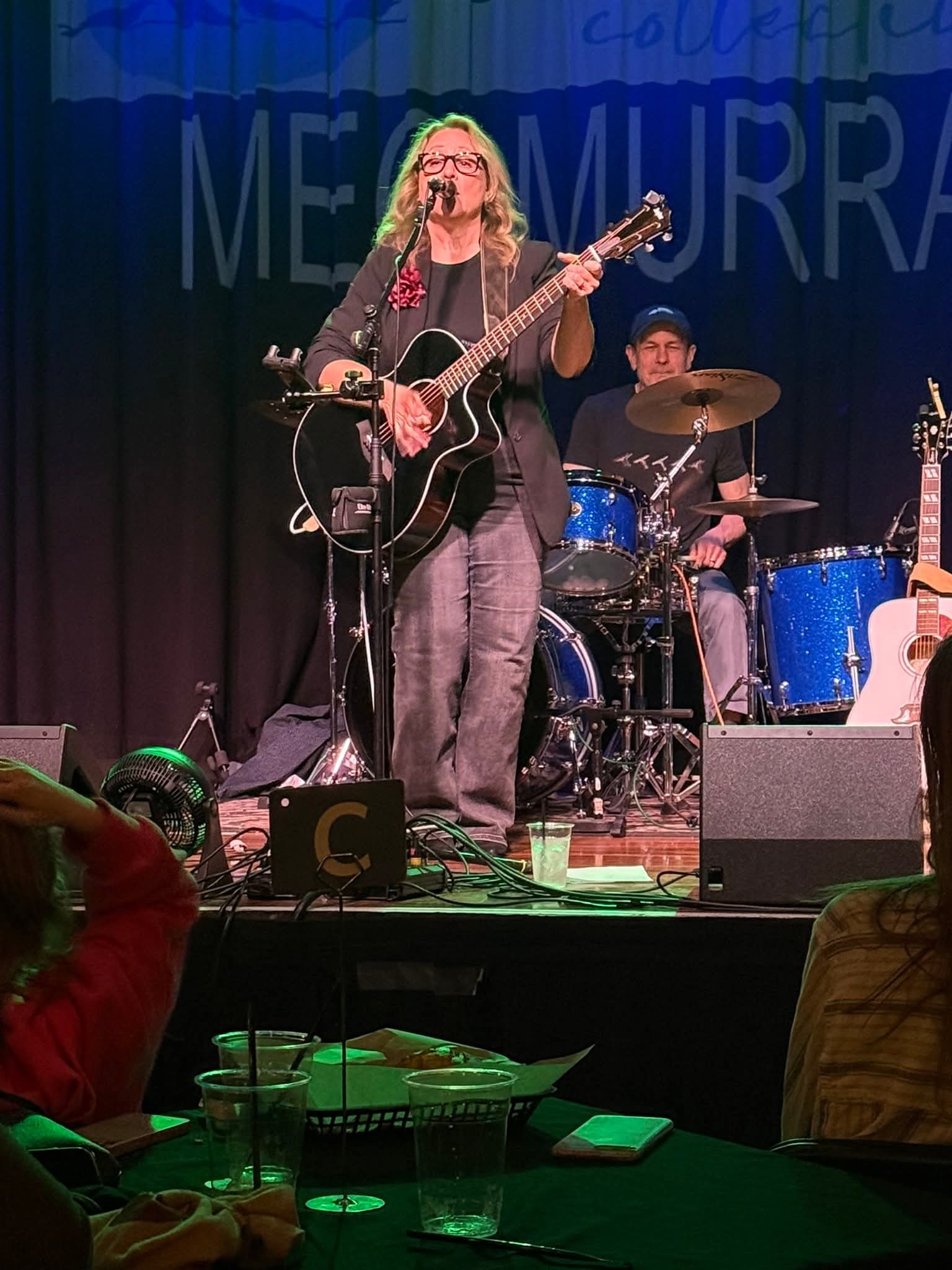 Musician with long hair playing guitar on stage, drummer behind, dark background, audience in foreground, stage lighting.