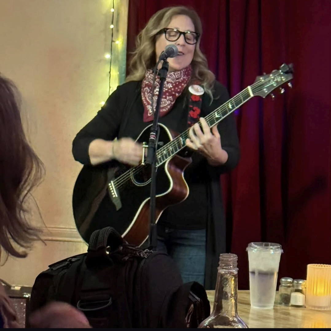 Woman with glasses singing and playing guitar on stage with red curtain background, microphone in front, drinks on table.