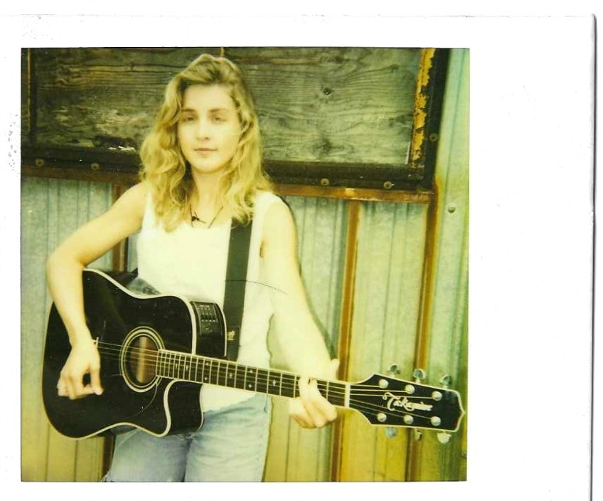 Young woman with wavy hair holding a black acoustic guitar, standing outdoors against a wooden and metal background.