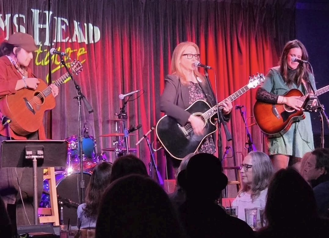 Three women perform on stage with guitars, in front of a red curtain, audience visible in foreground.