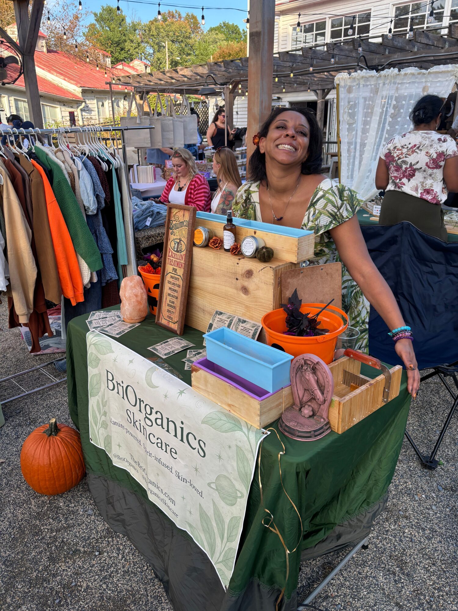 Woman at an outdoor market stall with organic products, pumpkins, and colorful items, smiling and waving. People and buildings in background.