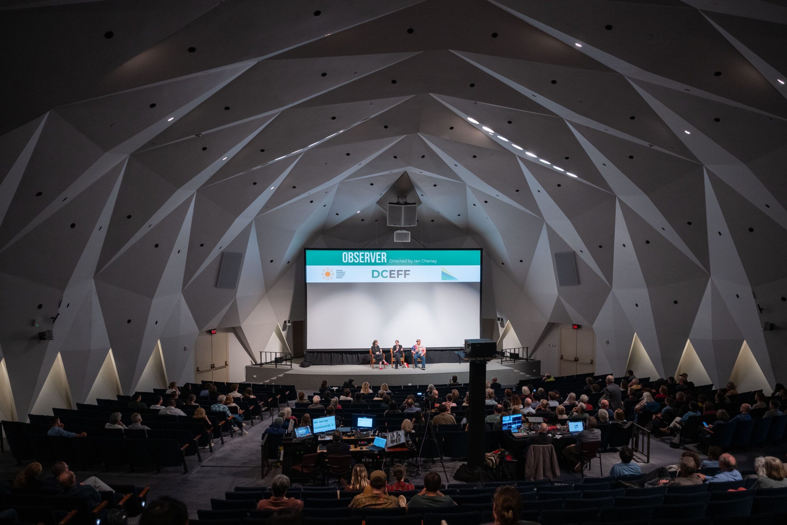 Conference auditorium with a stage, projection screen, seated audience, and geometric faceted ceiling.