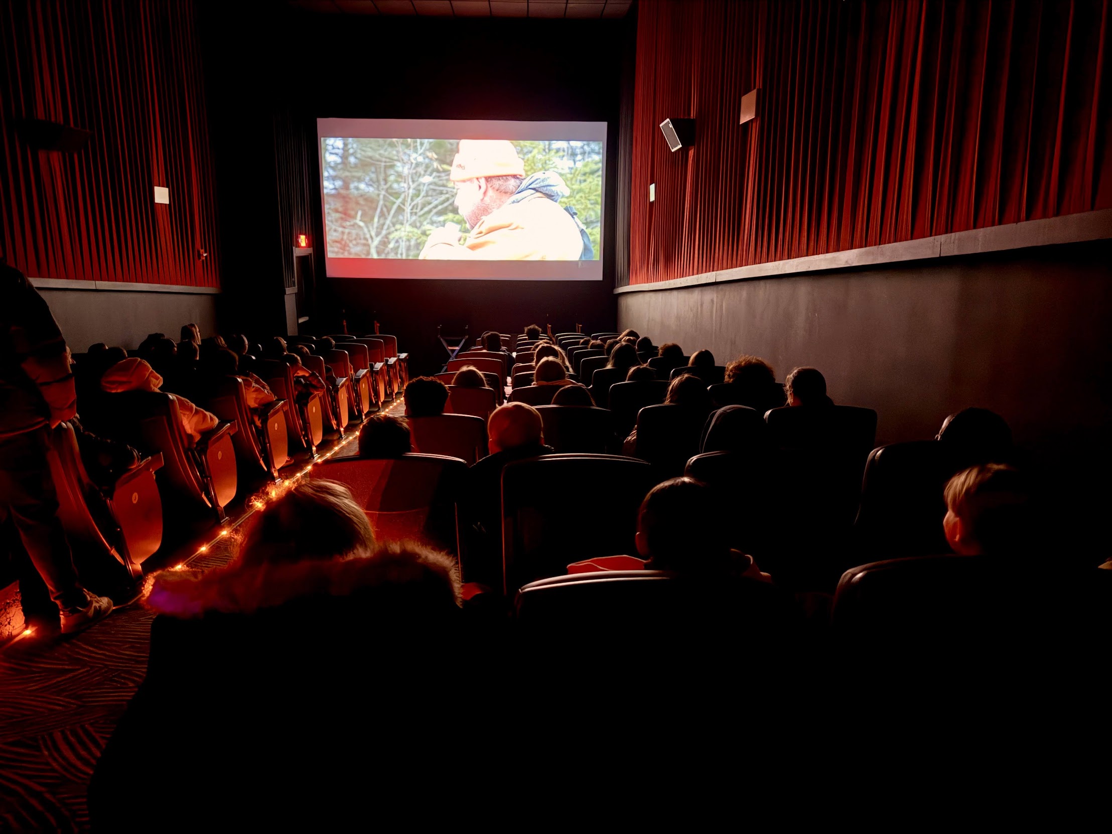 Audience seated in a red-lit theater watches a film on a large screen.