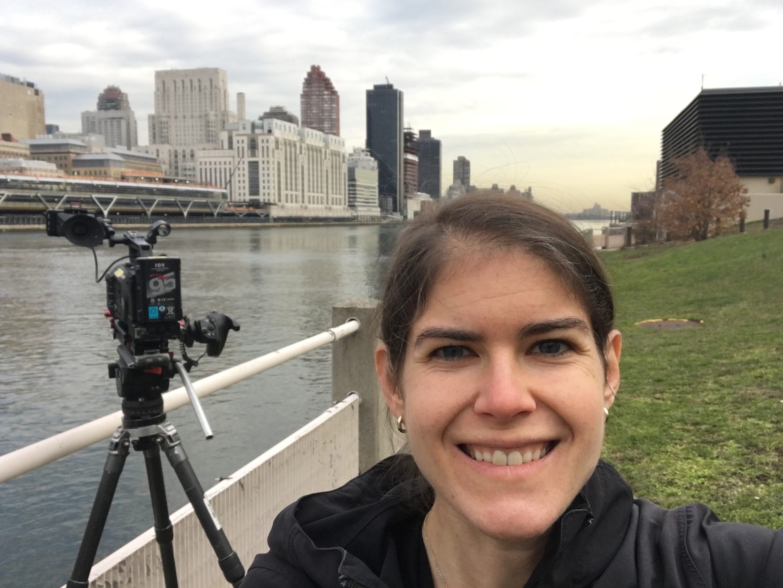 A person smiles beside a camera on a tripod along a waterfront, with city buildings across the river.