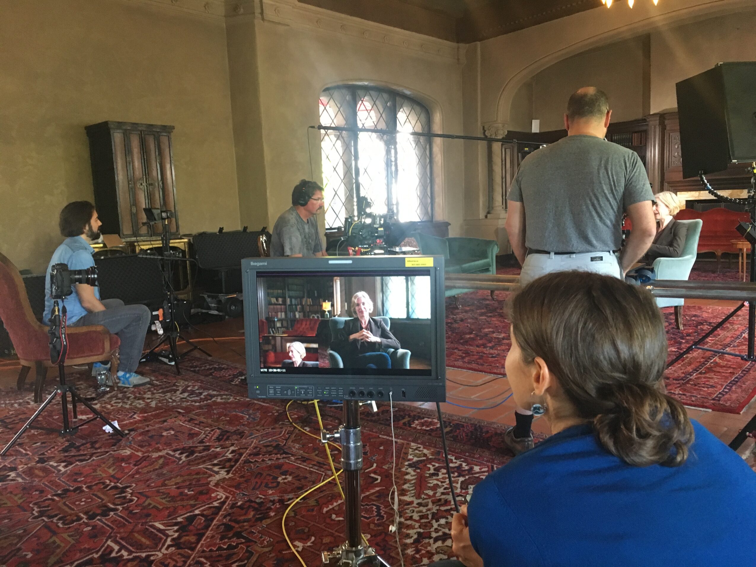 People sit in a room with a television on a stand, instruments, chairs, and patterned rugs.