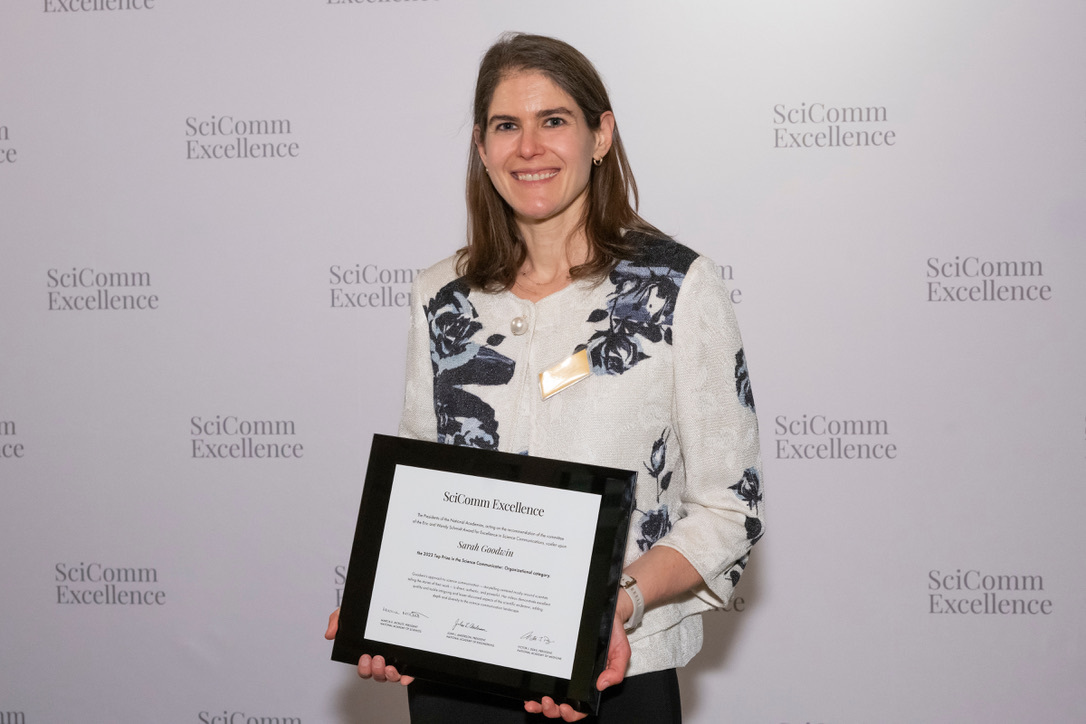 A person holds a framed certificate in front of a SciComm Excellence backdrop.
