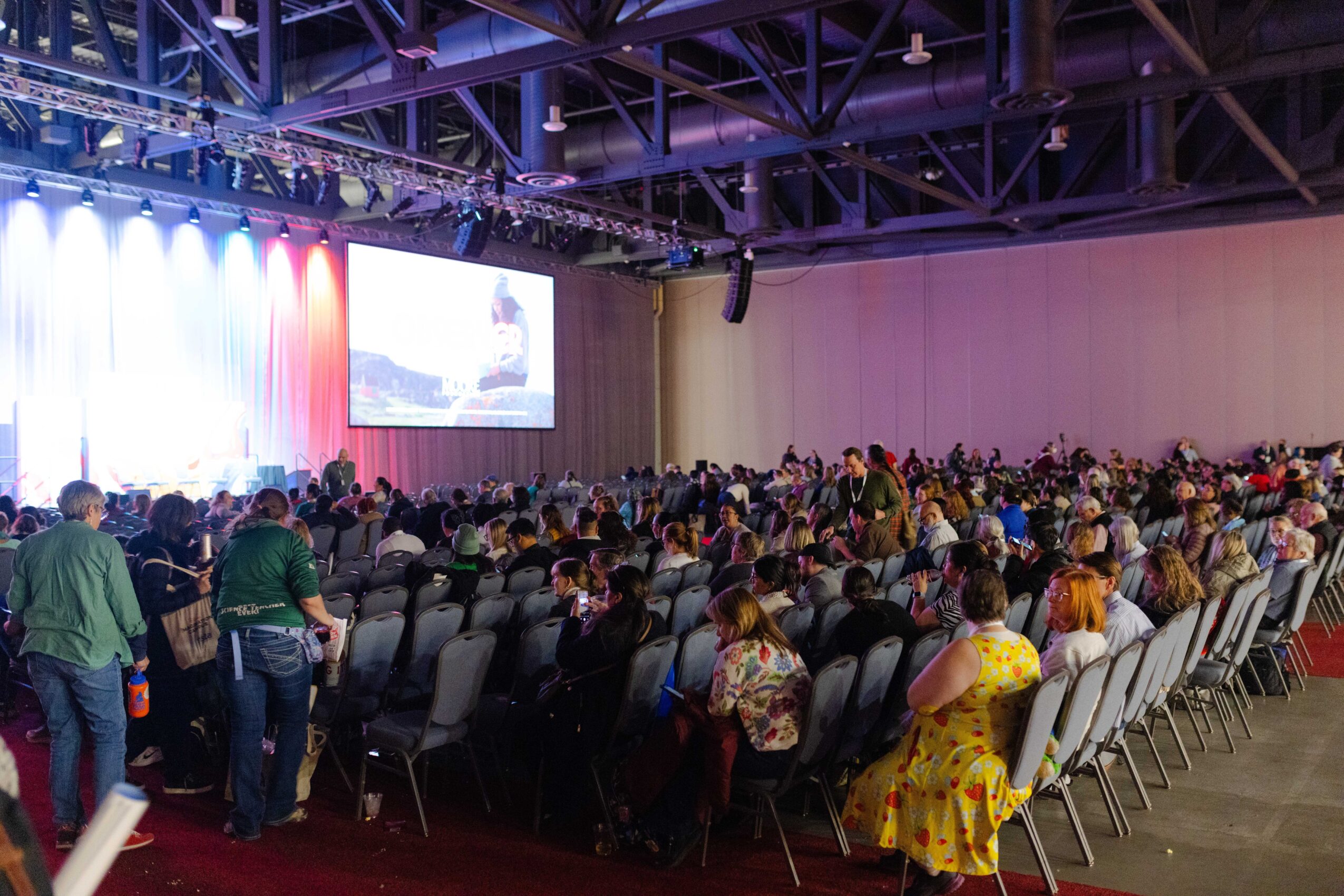 A large audience sits in rows facing a projection screen in an event hall with colorful stage lighting.