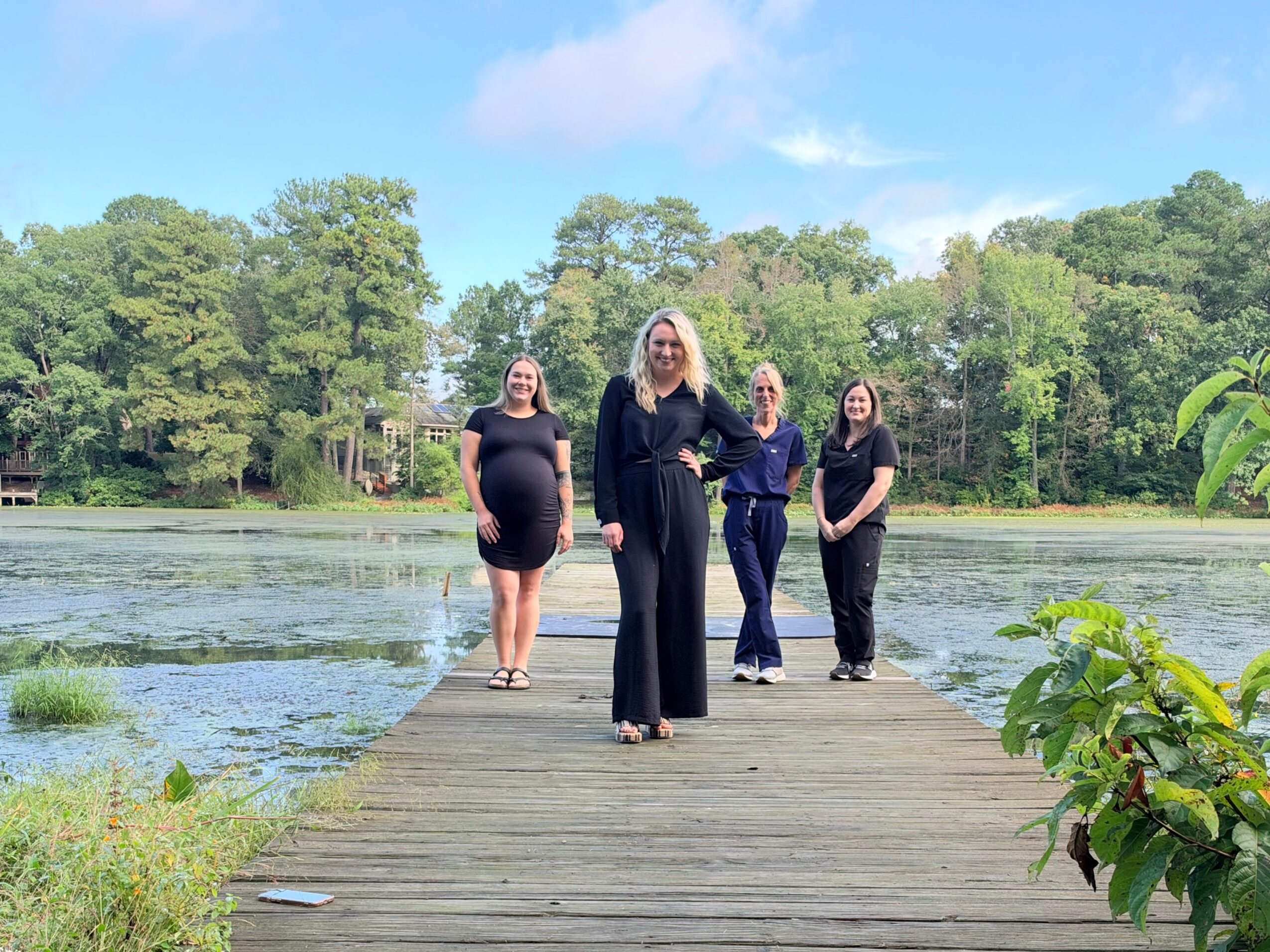 Four people stand on a wooden dock beside a pond, with trees and blue sky behind them.