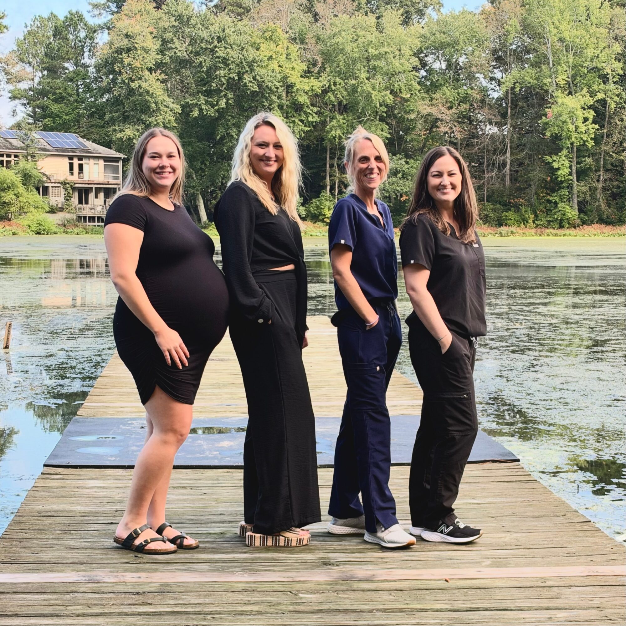 Four people standing on a wooden dock beside a pond, with trees and a building in the background.
