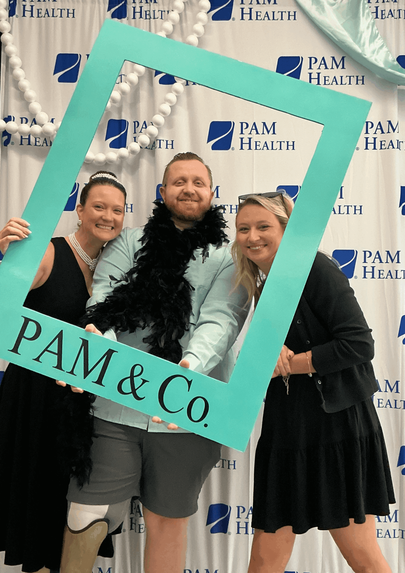 Three people pose inside a teal picture-frame prop in front of a PAM Health backdrop.