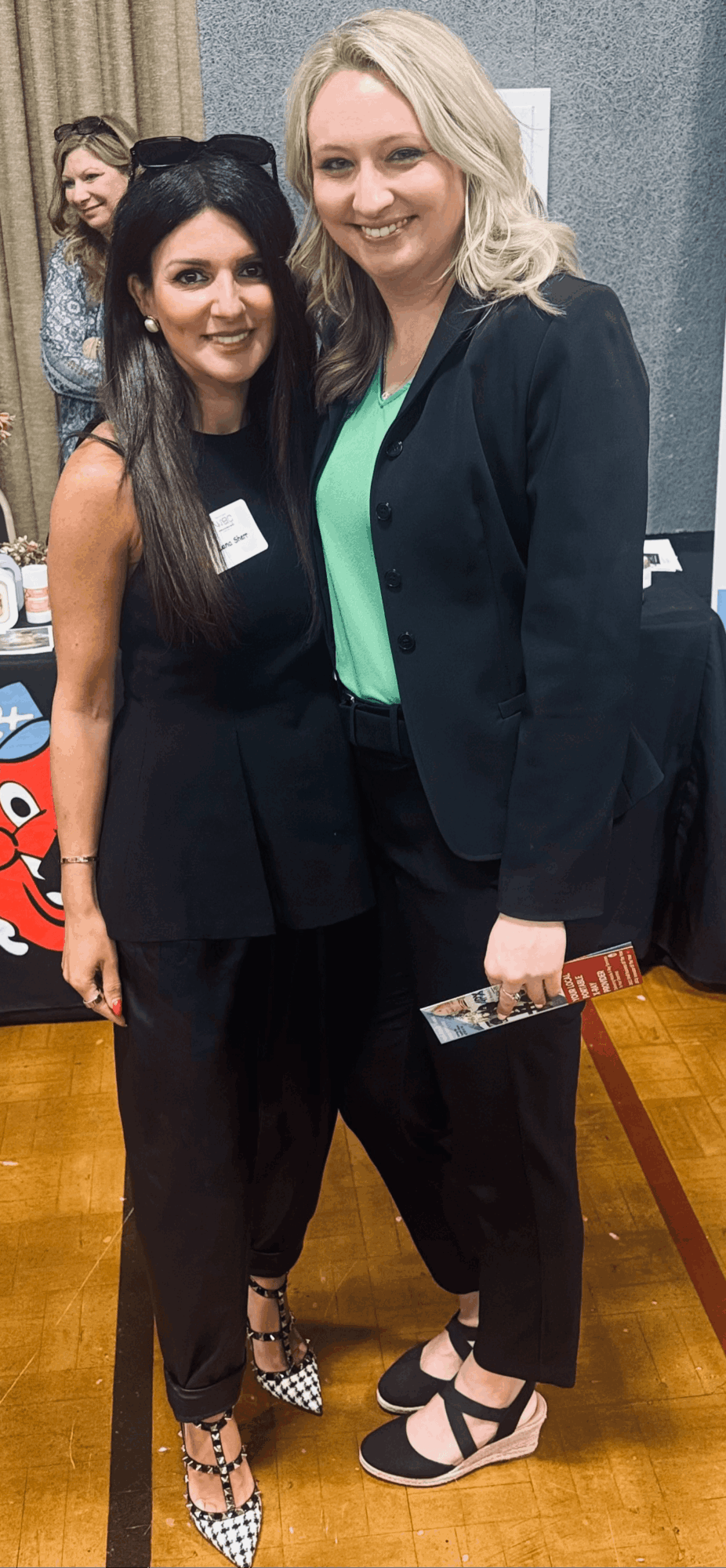 Two people pose together at an indoor event, with another person visible behind them.