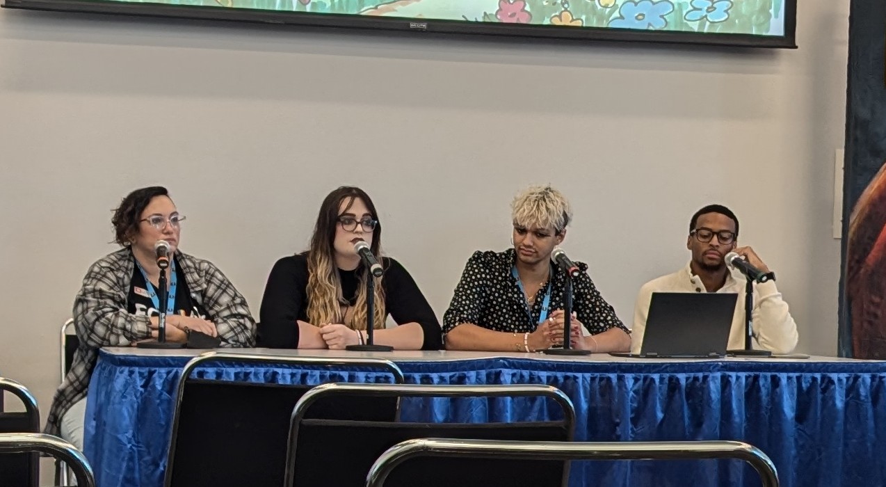 Four people sit behind a blue-draped panel table with microphones and a laptop.