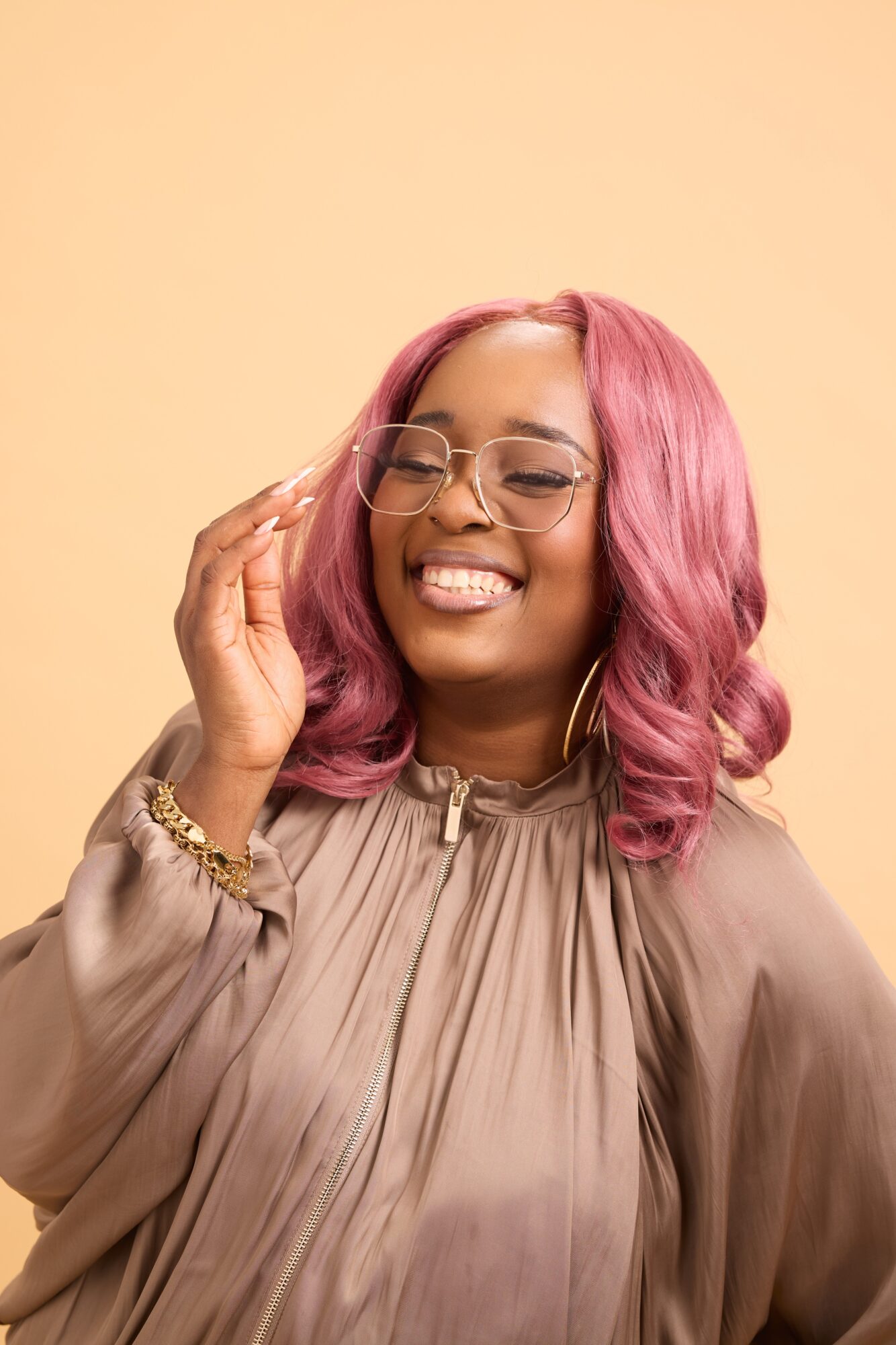 Person with pink wavy hair and glasses smiling while adjusting the frames, wearing a taupe blouse against a plain background.
