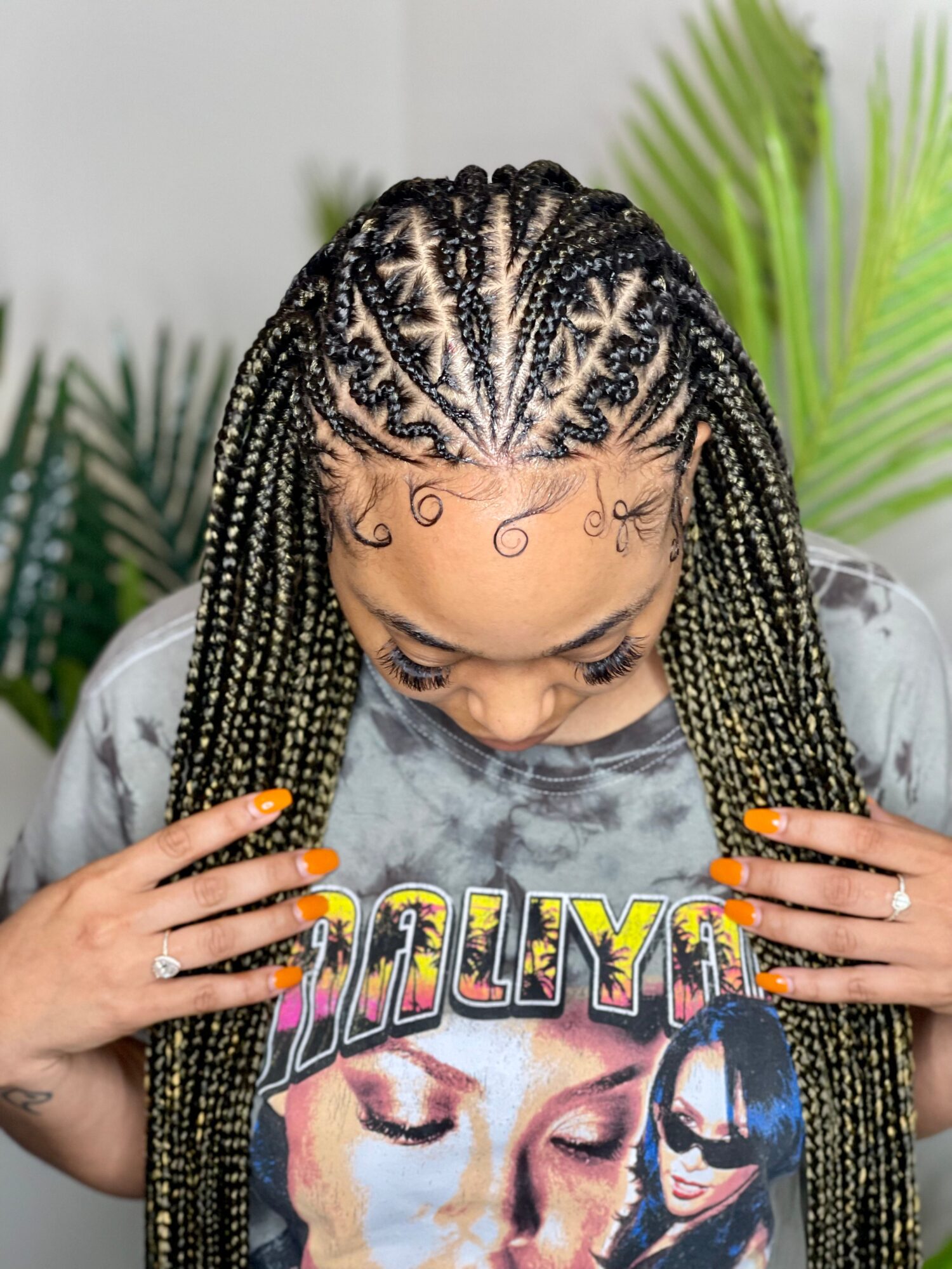 Person with long braided hair looking down, wearing a graphic T-shirt in front of leafy plants.
