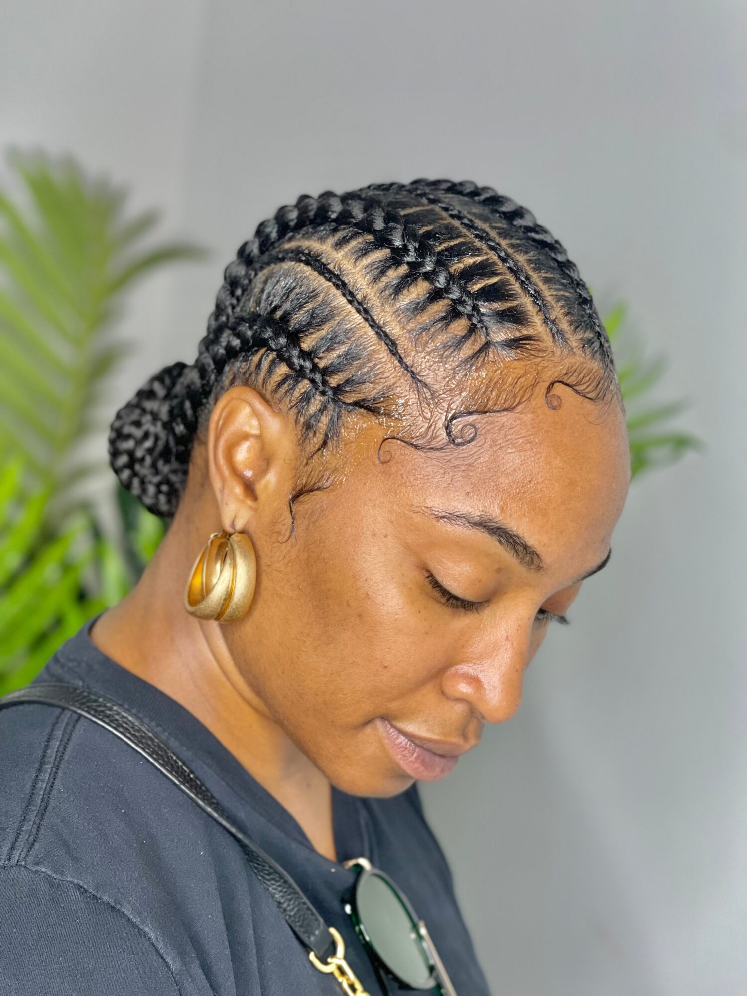 Person with braided hair and gold hoop earrings looking downward, wearing a dark top and sunglasses around the neck.