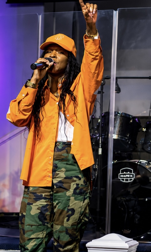 Performer in an orange jacket and camouflage pants sings into a microphone with one arm raised onstage.