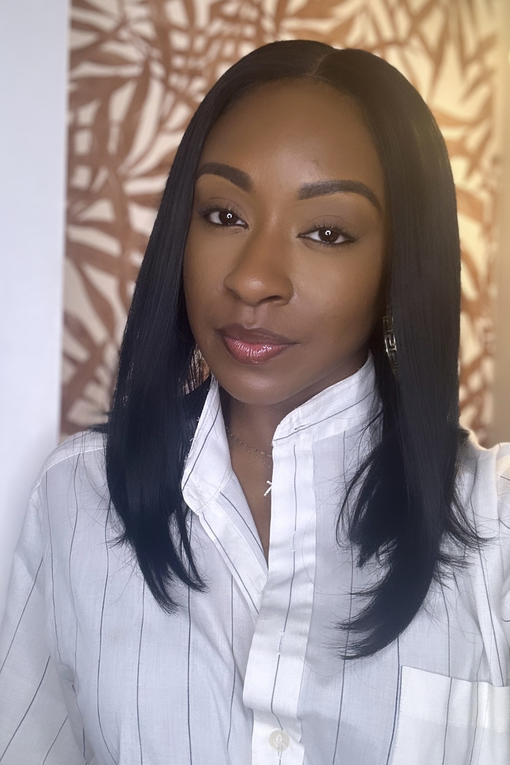 Person with long dark hair wearing a white pinstriped shirt, standing before a patterned wall.