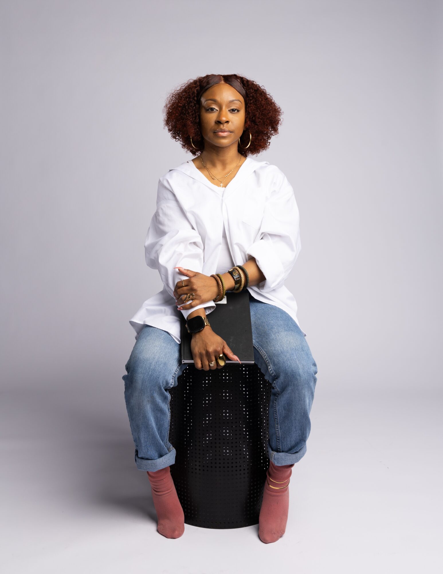 A seated person in a white shirt and jeans holds a black bag against a gray studio background.