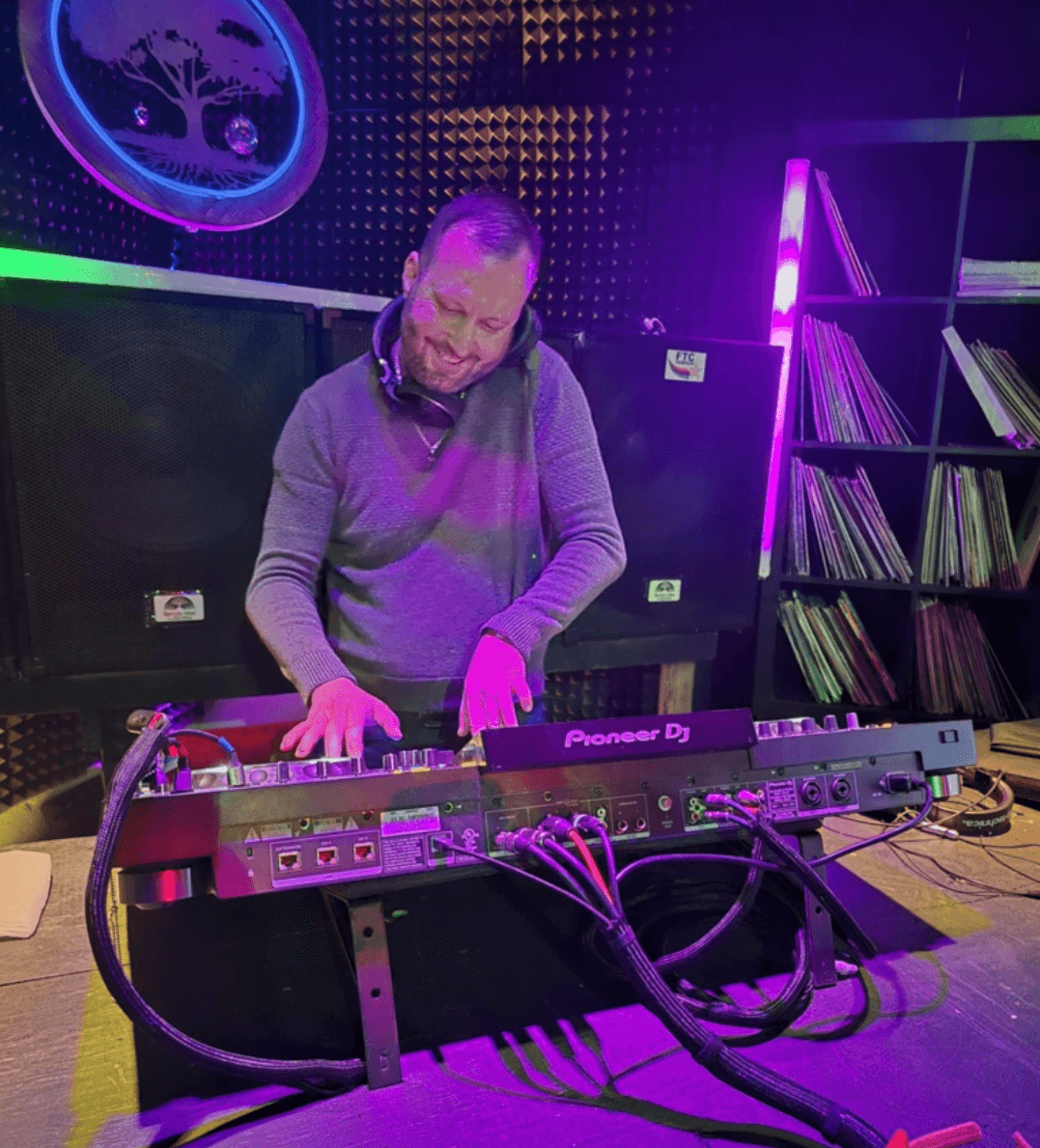 Person operating a Pioneer DJ controller in a neon-lit room with records on shelves.