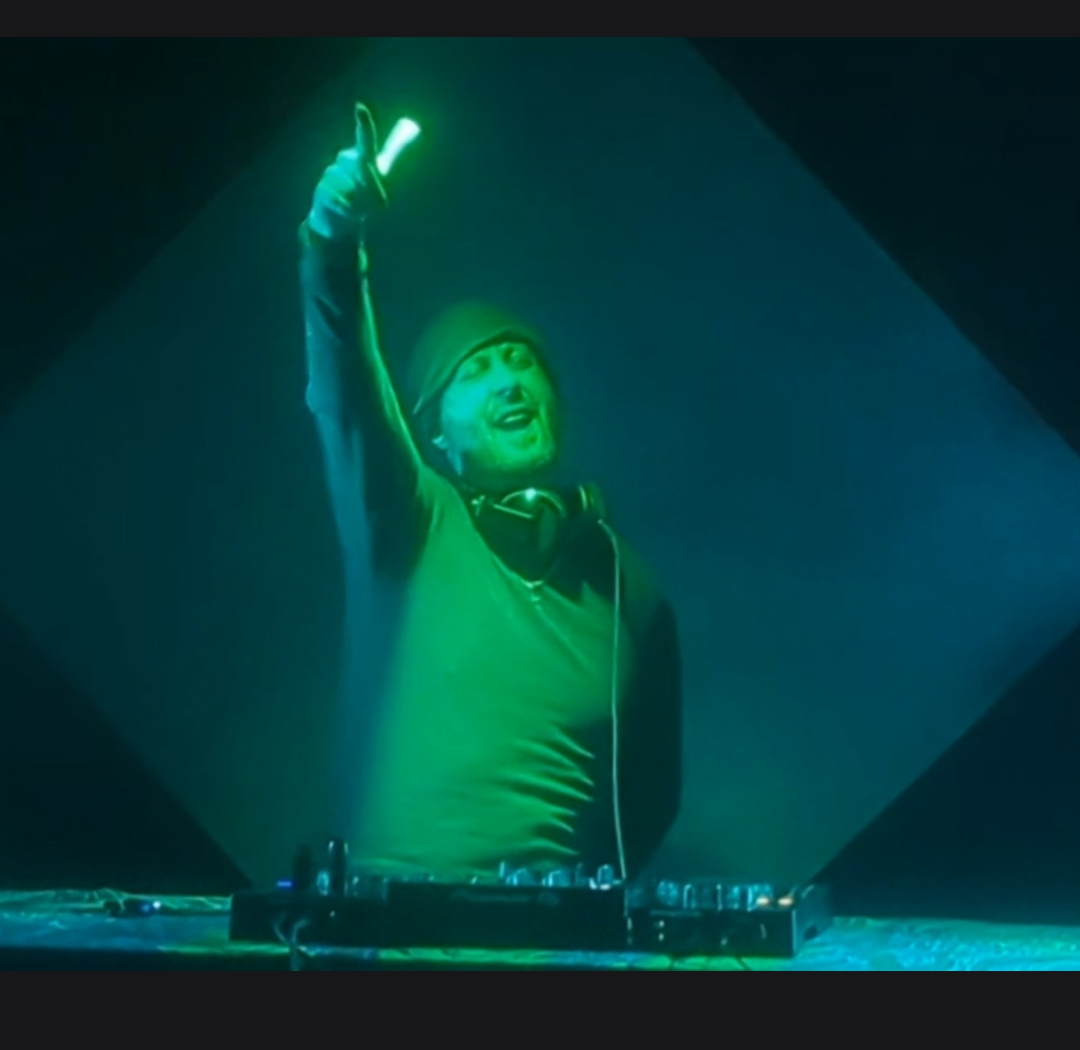 A DJ raises one hand while standing behind a mixing console under green stage lighting.