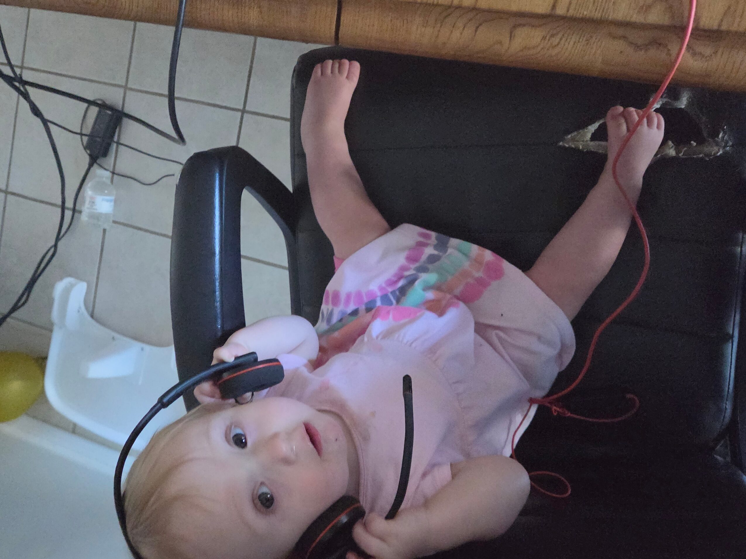 A baby lies on a black chair beside a wooden table, with cables nearby.