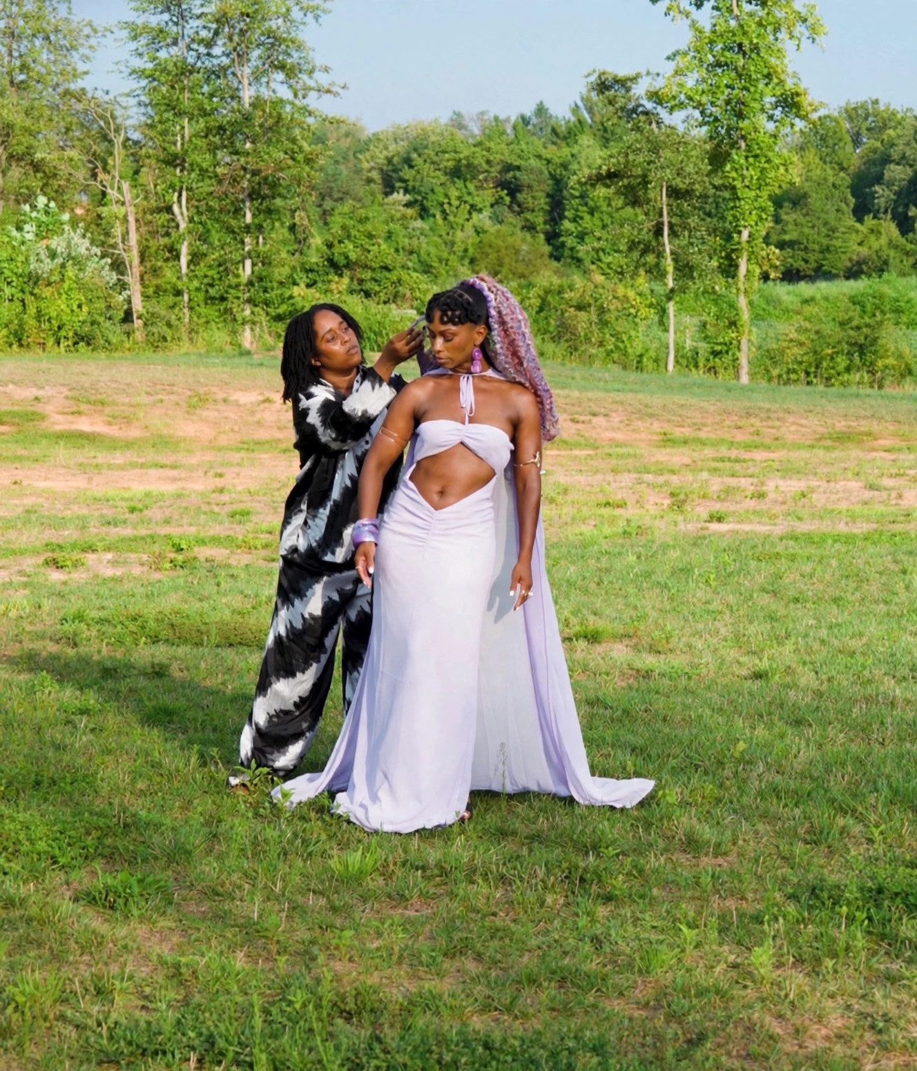 Two people stand on a grassy field; one adjusts the other’s veil as they wear a white gown.