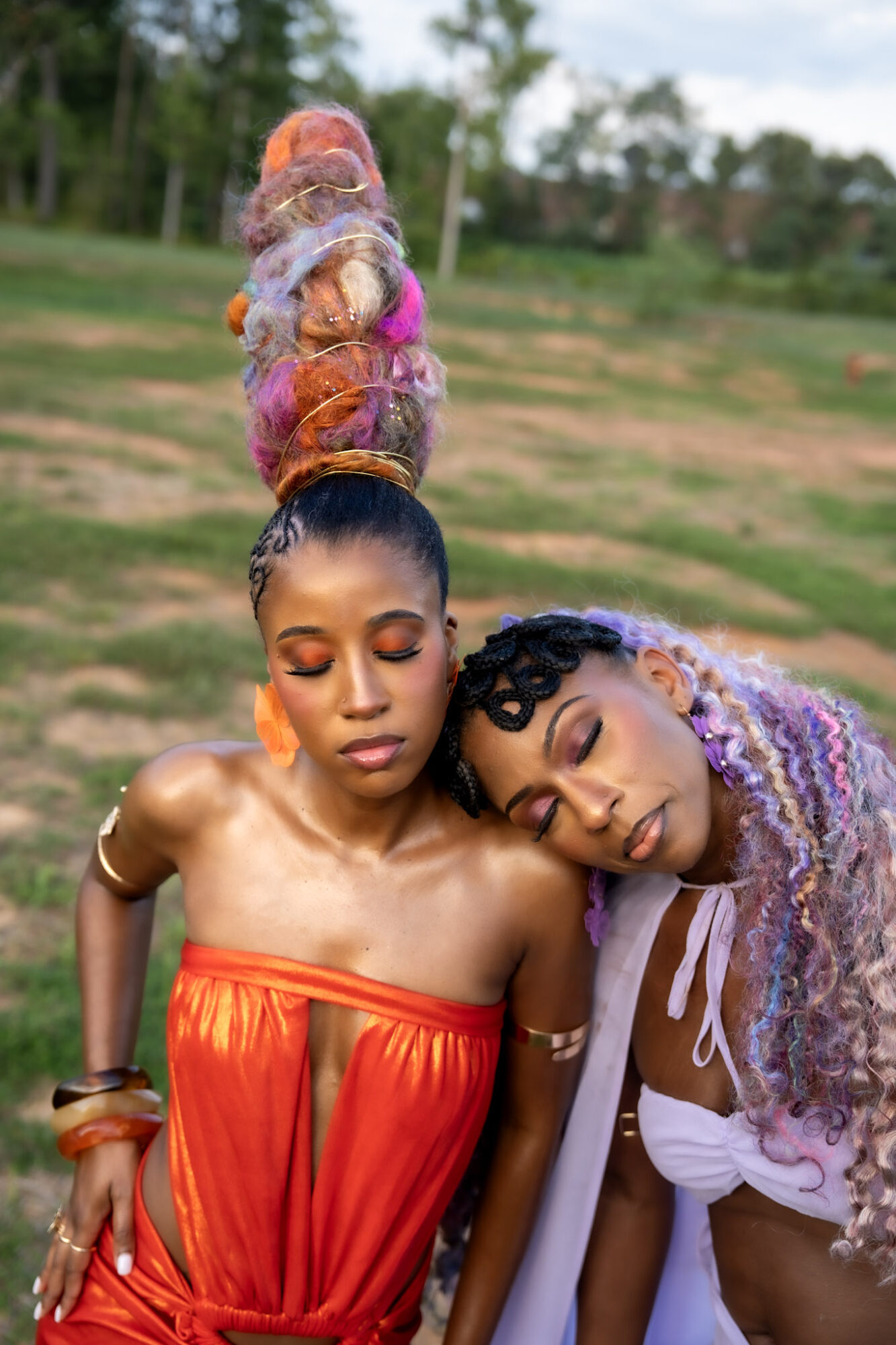 Two people lean together outdoors; one wears a red dress and an elaborate high bun, the other has long lavender braids and a white top.