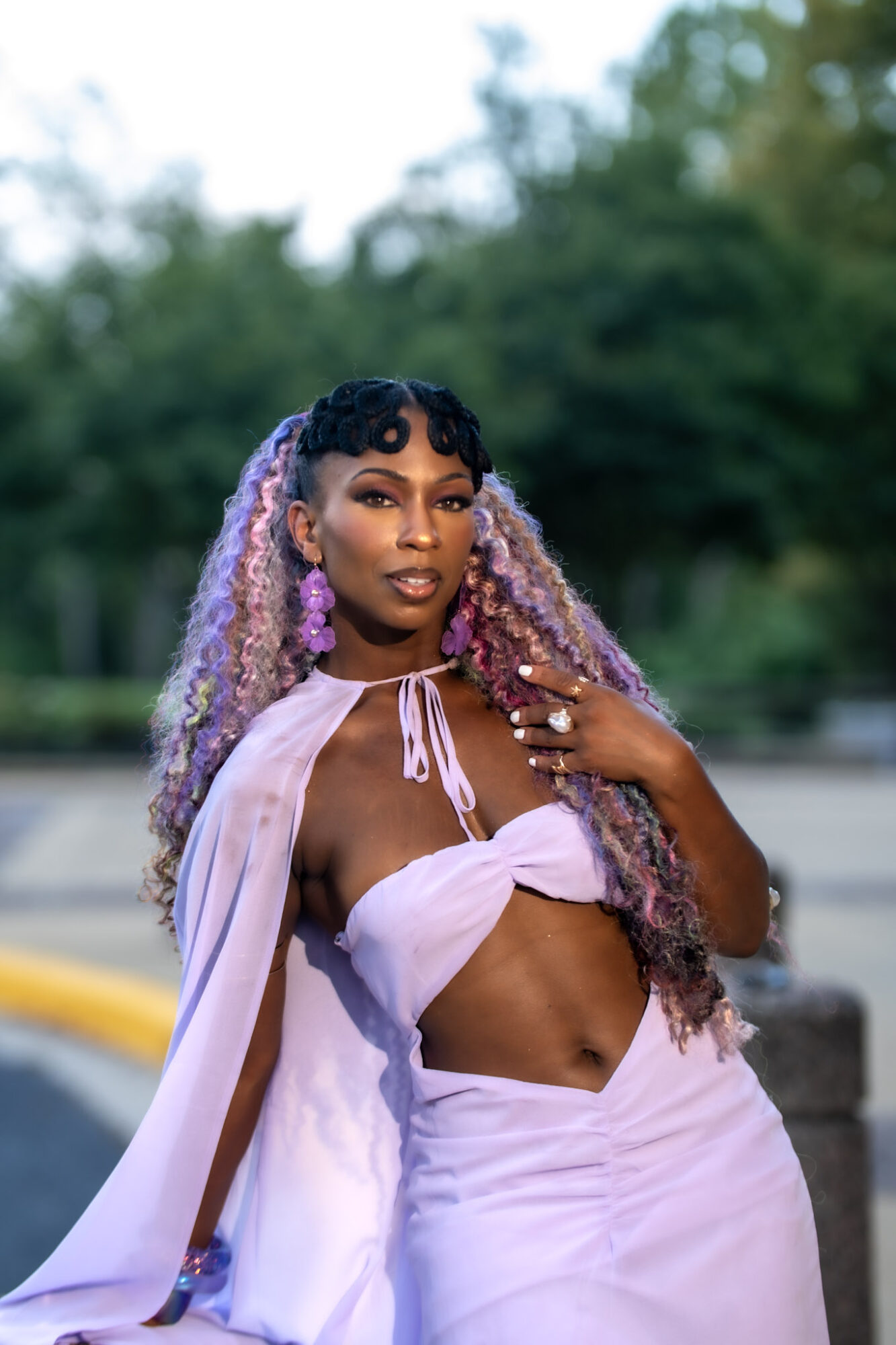Person with long purple hair wearing a pale pink outfit, standing outdoors near greenery and pavement.