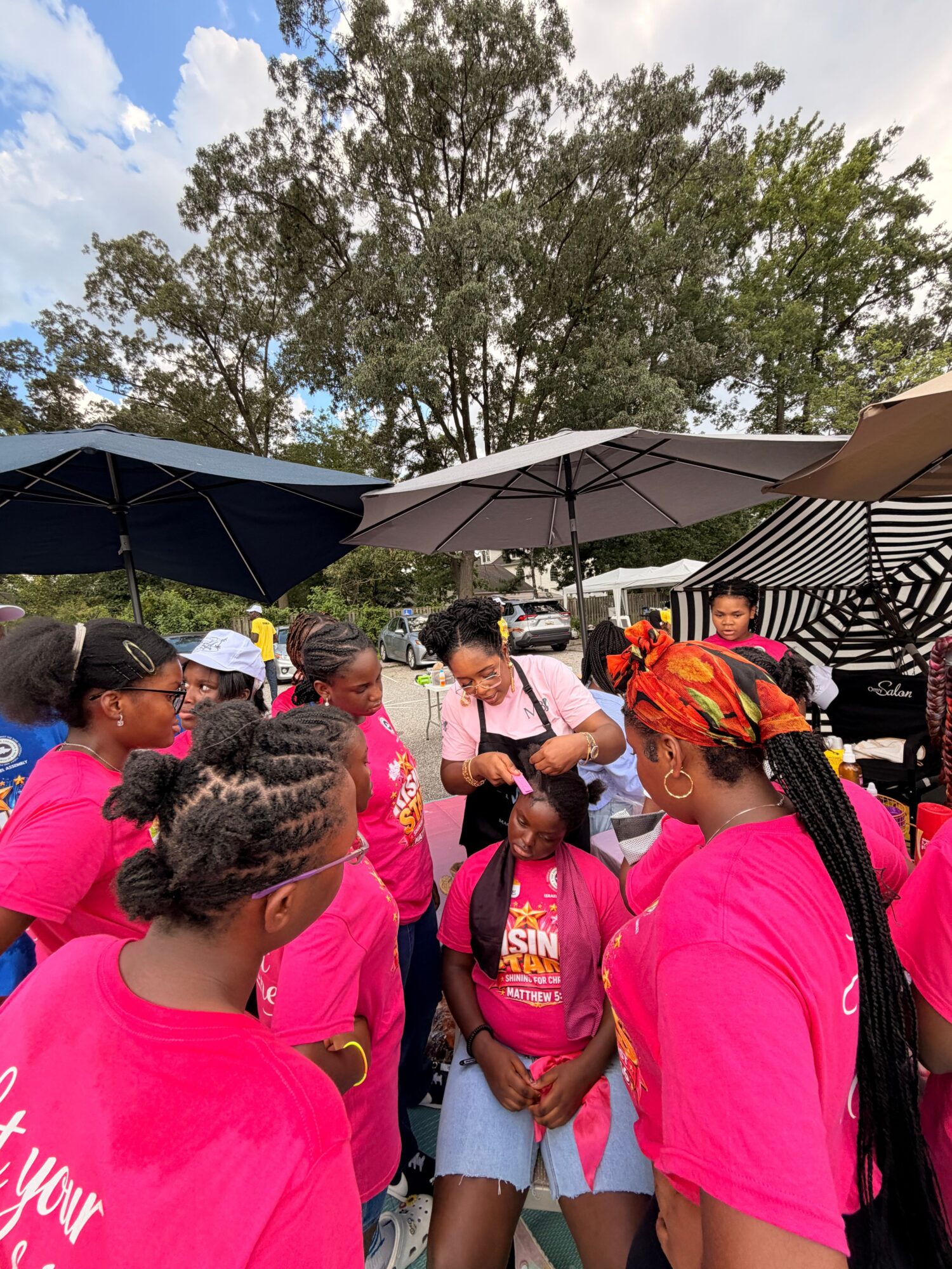 People in bright pink shirts gather beneath canopies outdoors near a large tree.