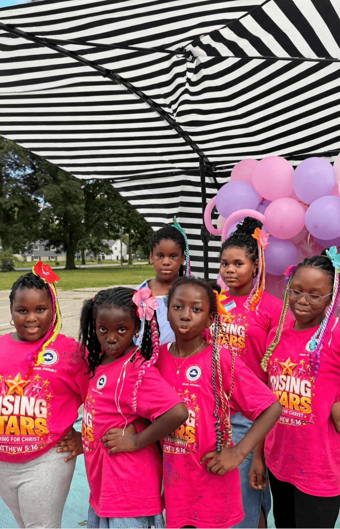Five people in bright pink shirts stand beneath a striped canopy beside purple and pink balloons.