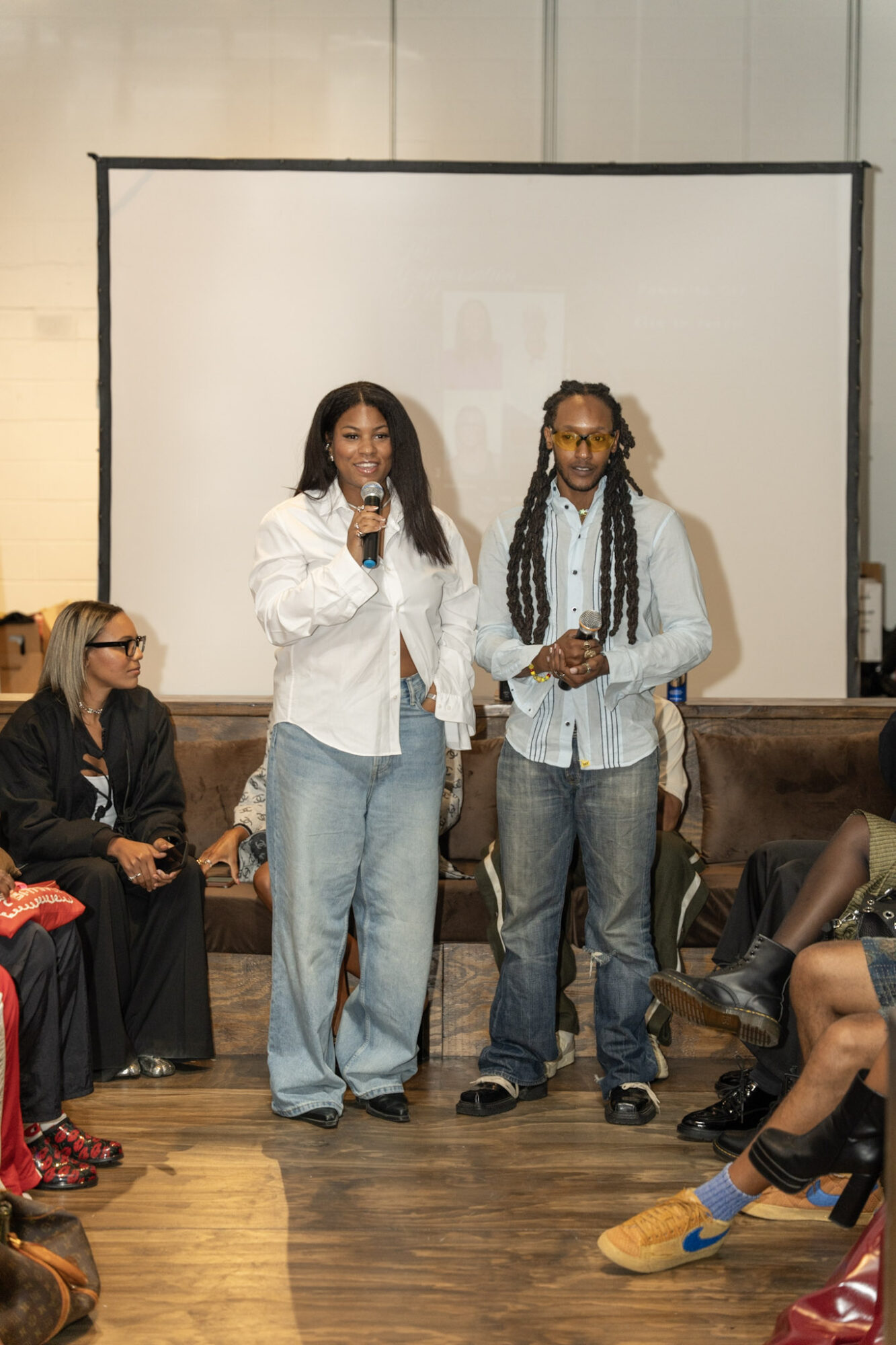 Two people stand in front of a projection screen; one holds a microphone, while seated people watch nearby.